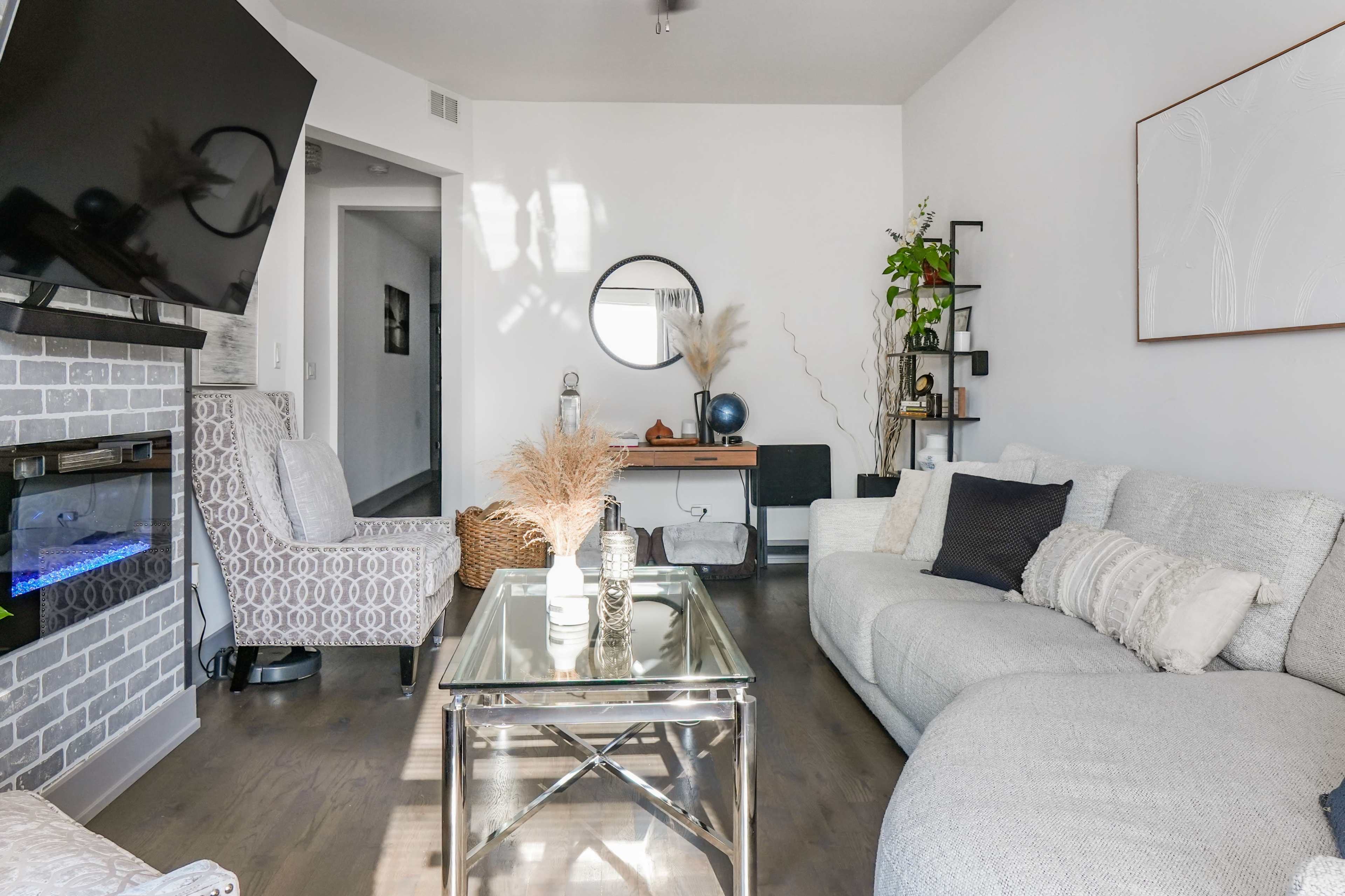 The image shows a modern living room featuring a light gray sectional sofa, a glass coffee table, a wall-mounted television, and decorative plants.
