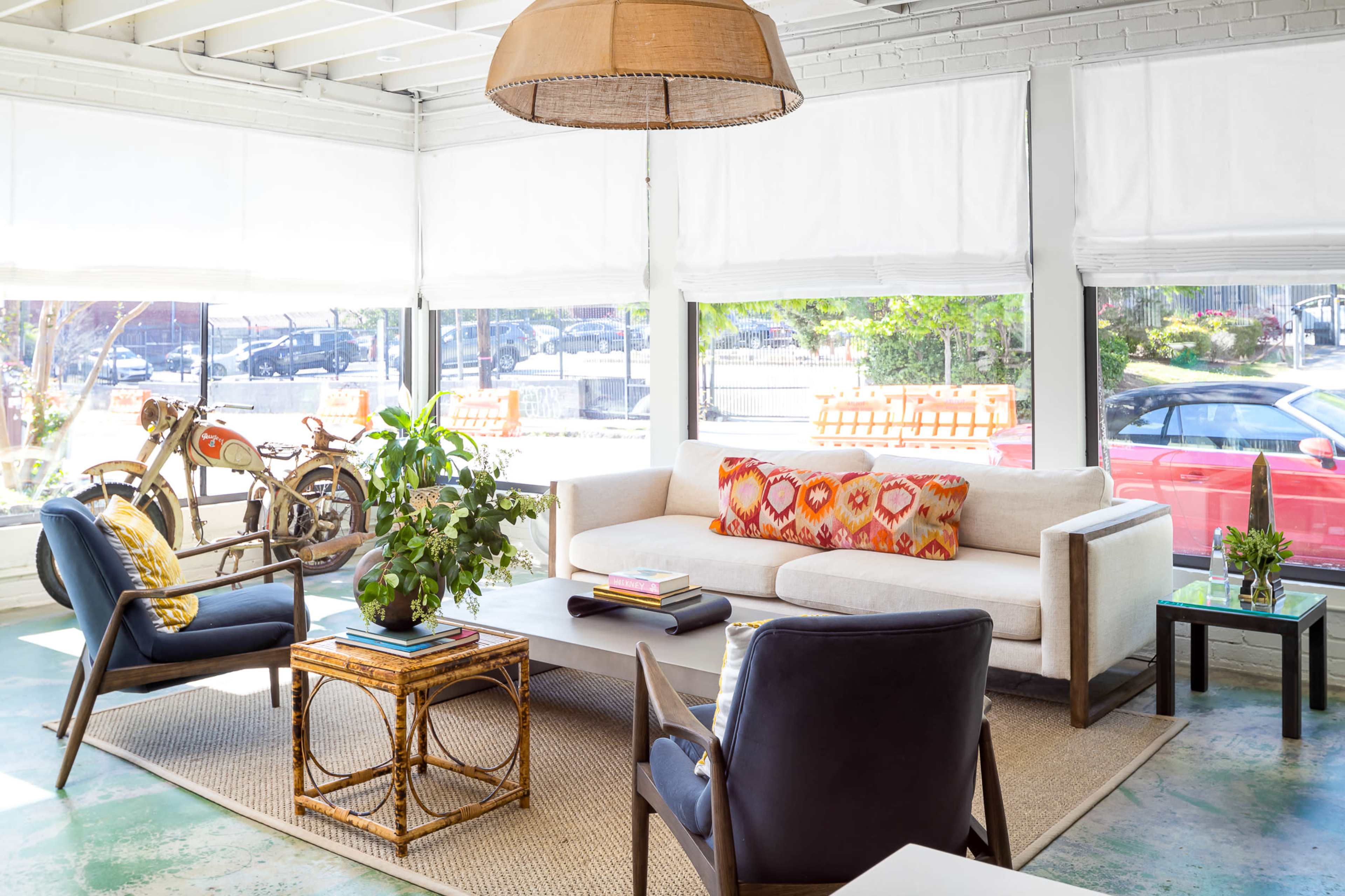 A bright living space featuring a beige sofa with a colorful cushion, a coffee table, a potted plant, and large windows with natural light, alongside a bicycle and a parked car visible outside.