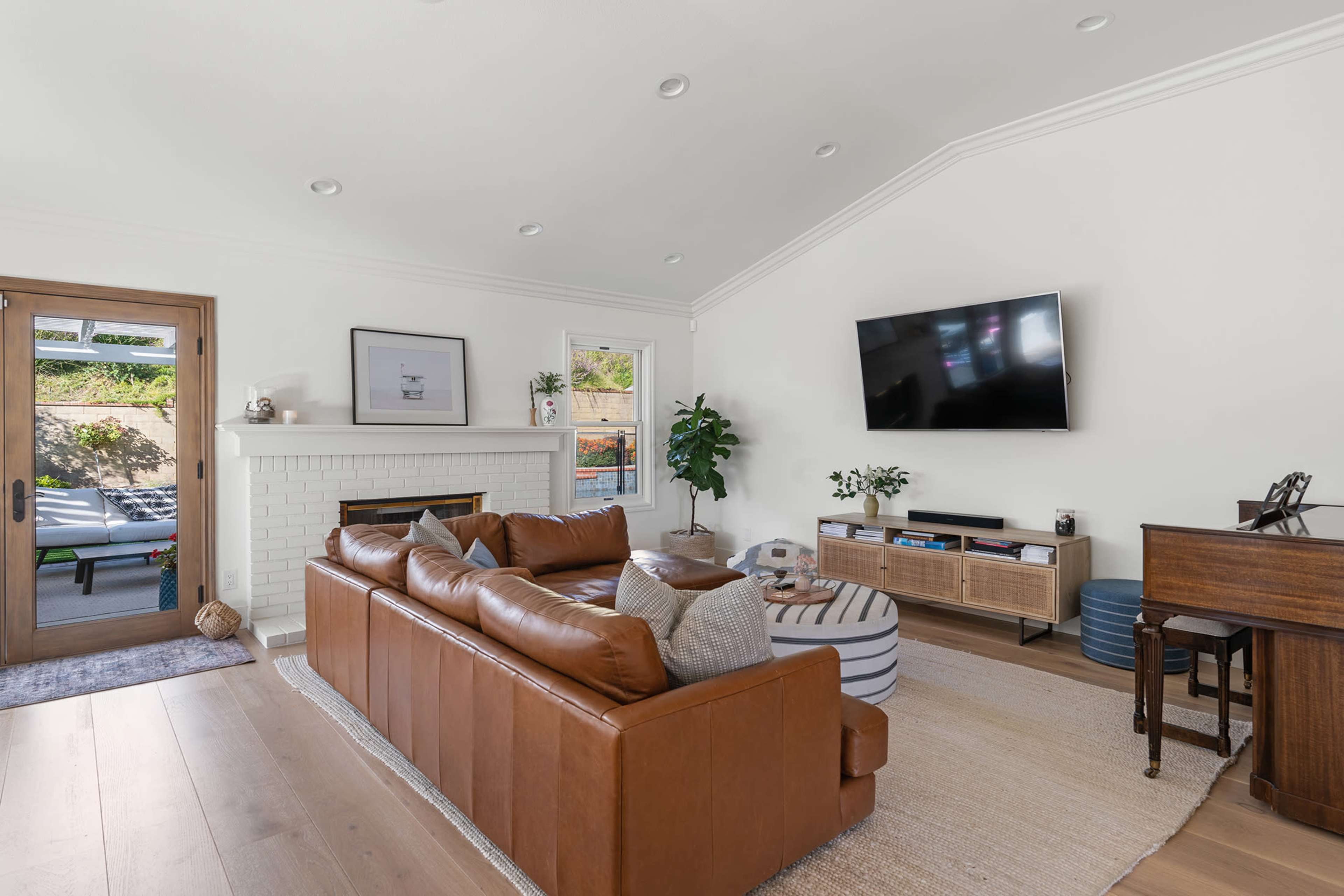 A modern living room features a brown leather sectional sofa, a white brick fireplace, a mounted television, and a mix of decorative and functional furniture.