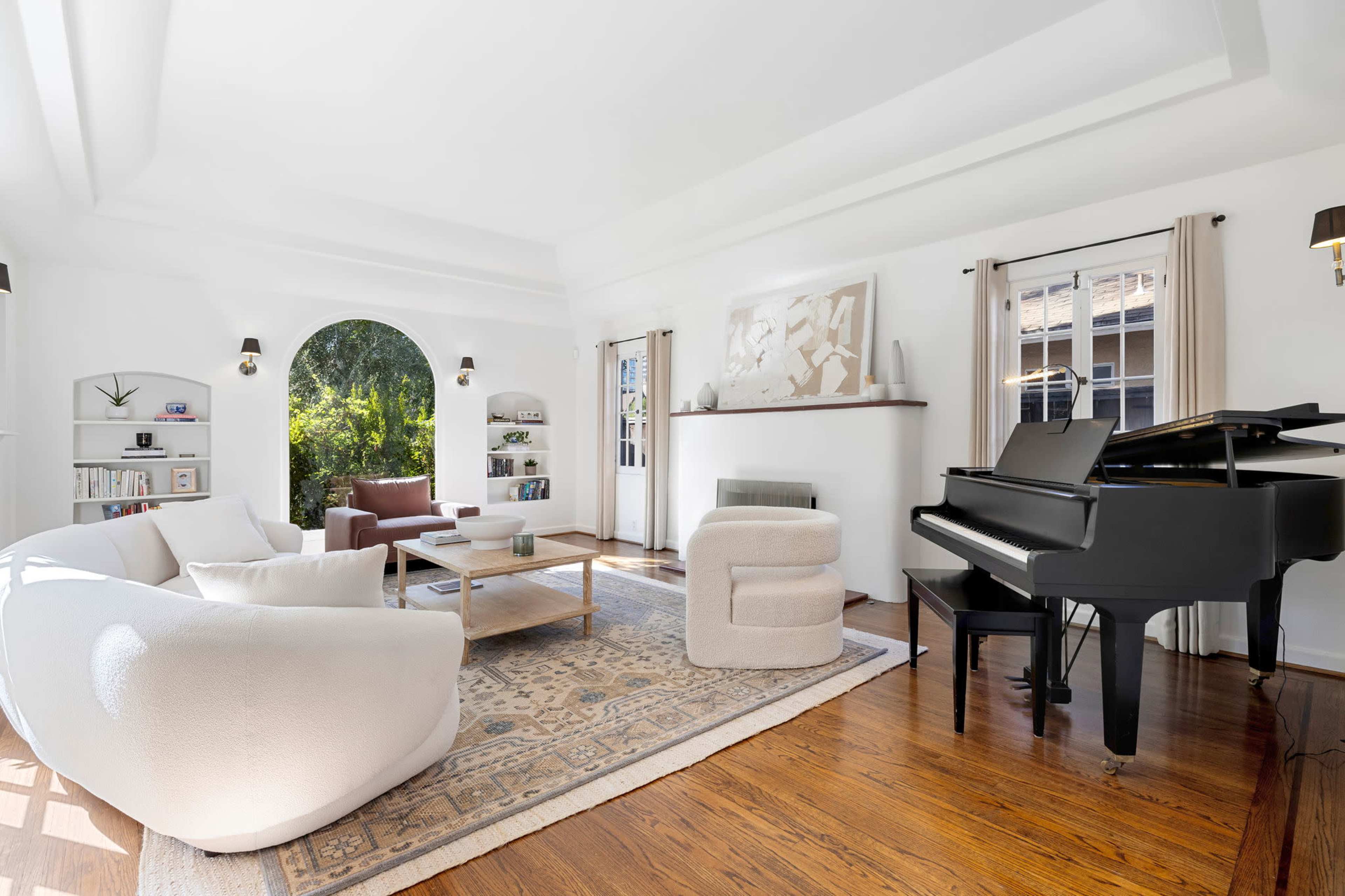 A bright living room features a white sofa and armchair, a wooden coffee table, and a black piano, with large windows allowing natural light.