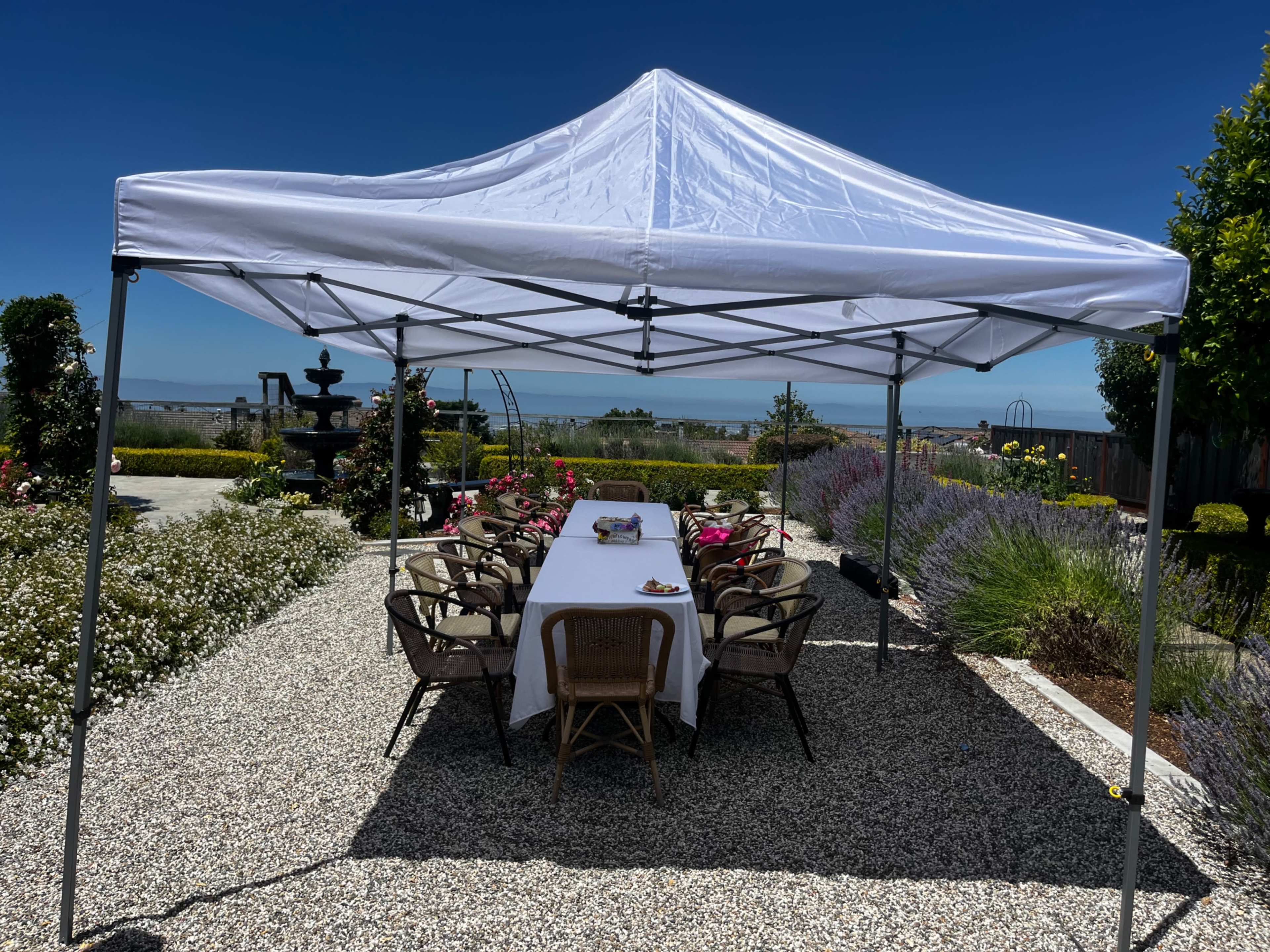 A white canopy covers a long table set for a meal, surrounded by chairs in a garden with flowers and a view of the ocean.