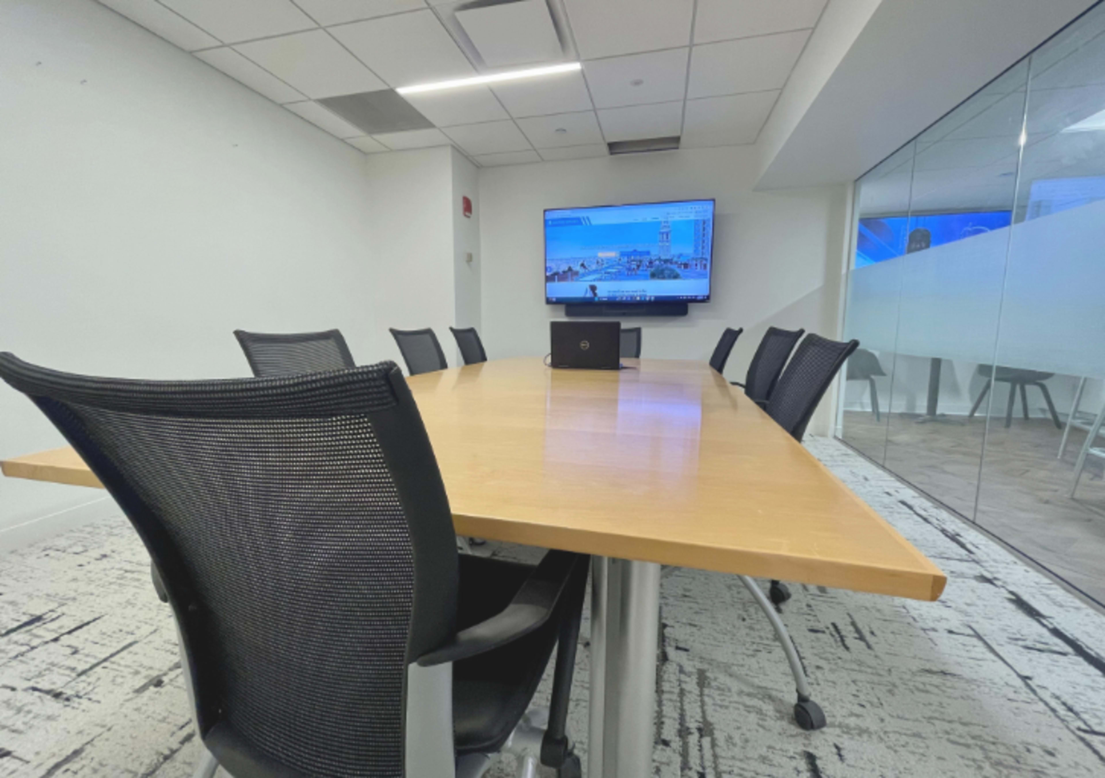 A long wooden conference table with black mesh chairs is positioned in a modern meeting room featuring a large screen mounted on the wall.