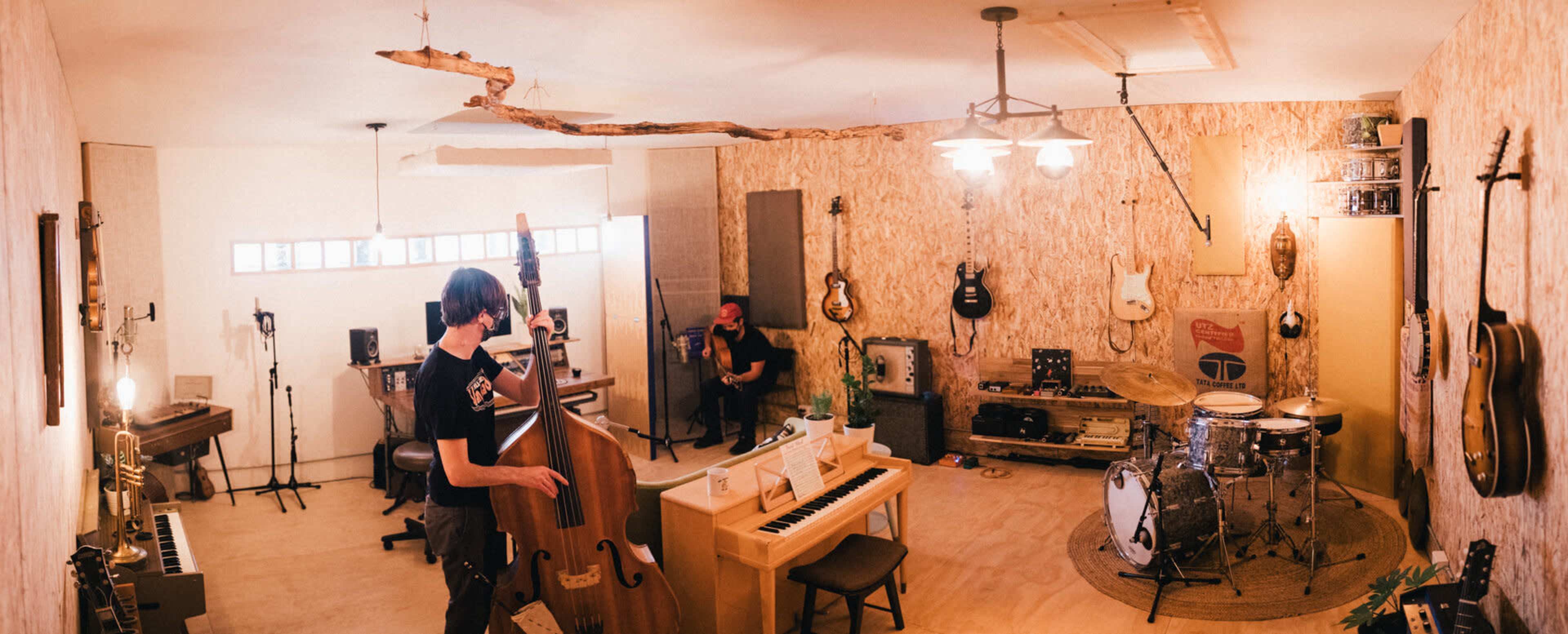 The image shows a music studio with a drummer, a bassist, and various musical instruments including a piano and electric guitars arranged in a well-lit, inviting space.