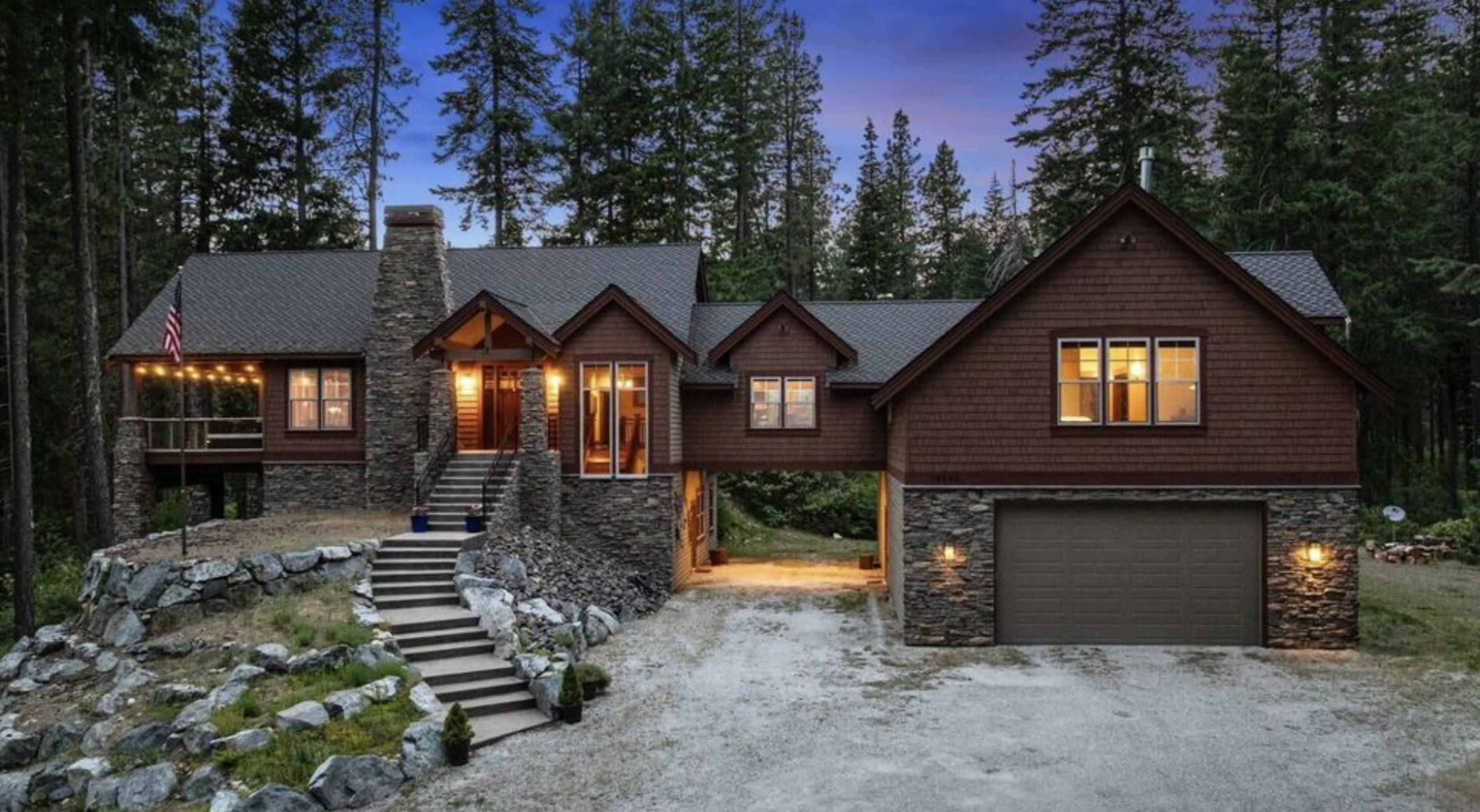 A large, rustic-style house with a stone facade and wooden accents is set amidst tall trees, featuring a three-car garage and a front porch with a staircase leading up to the entrance.