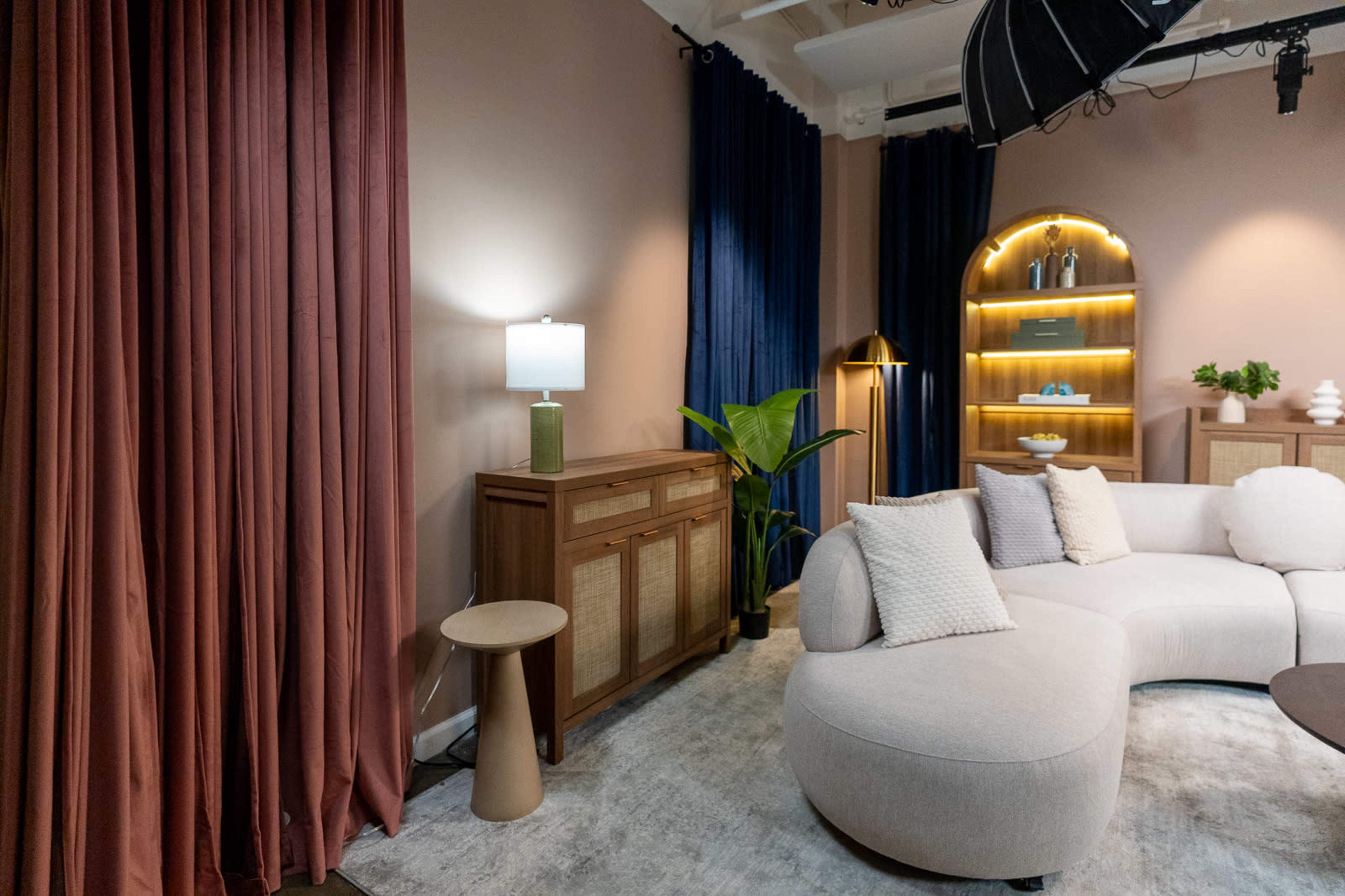 The image shows a modern, softly lit living room featuring a curved sofa, a wooden sideboard, a floor lamp, and decorative plants, all set against a backdrop of textured curtains.