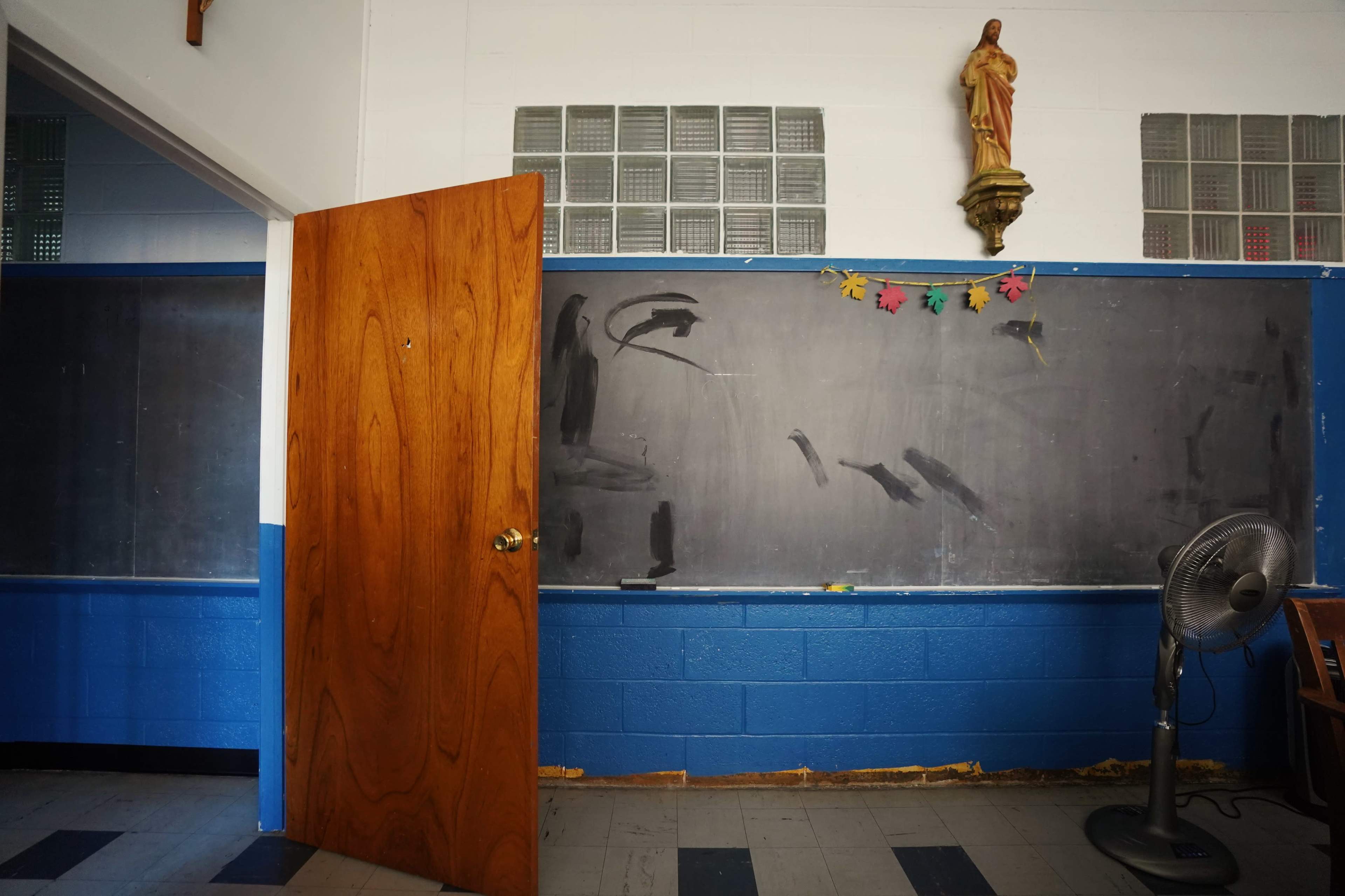 A classroom interior with a wooden door, a black chalkboard with some markings, and a fan near the wall.