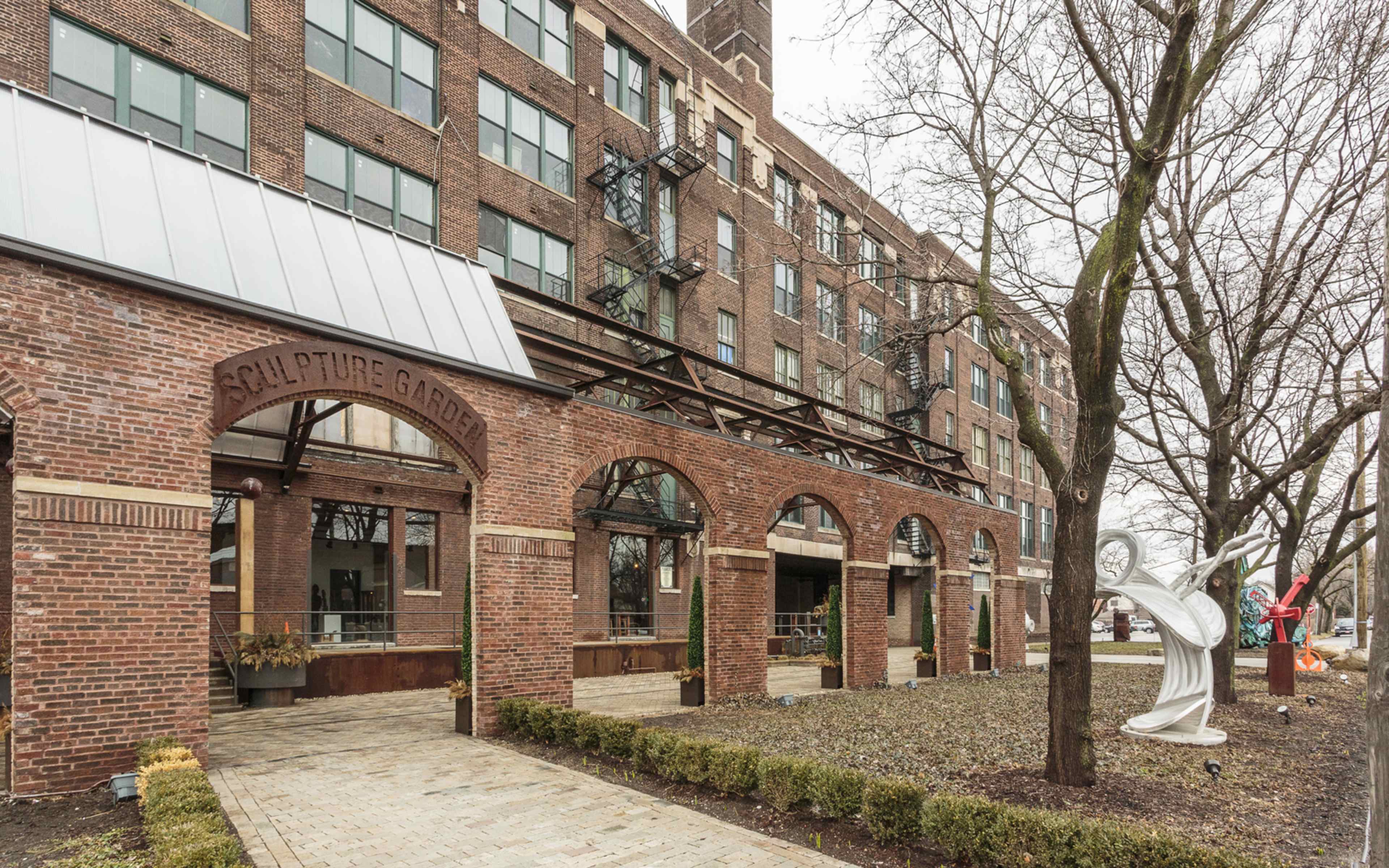 The image shows an entrance to a brick building featuring arches labeled "Sculpture Garden," with sculptures and greenery in the surrounding area.