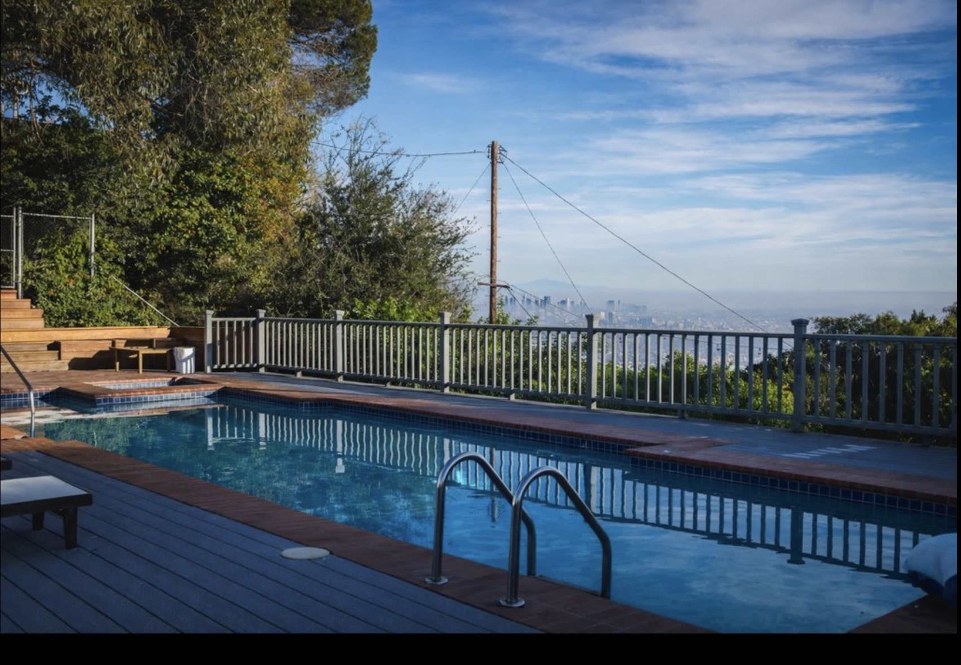 A swimming pool with metal ladders is surrounded by wooden decking and overlooks a cityscape under a clear blue sky.