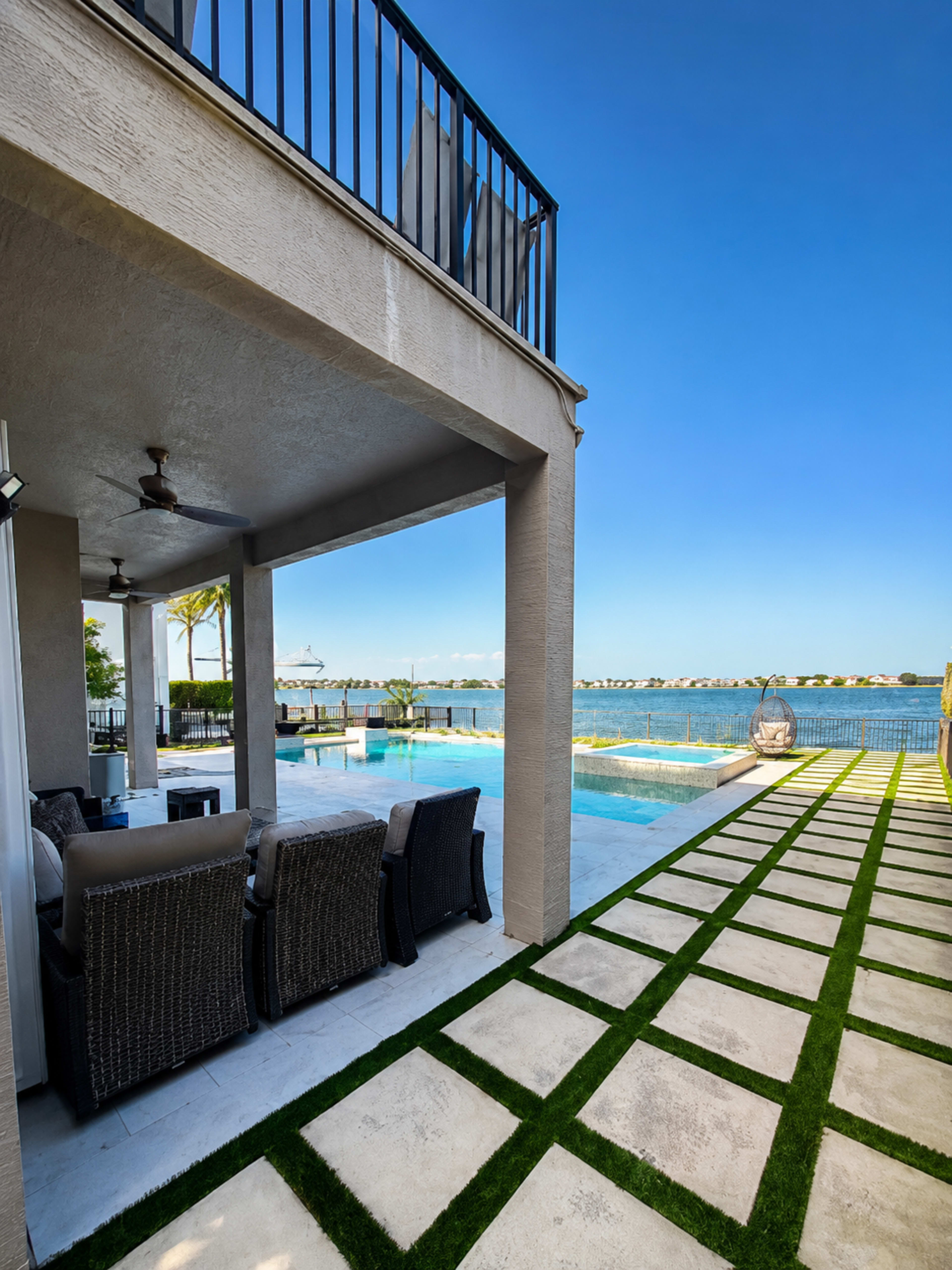 The image shows a waterfront property with a pool, a patio area furnished with seating, and a walkway lined with grass and stone tiles.