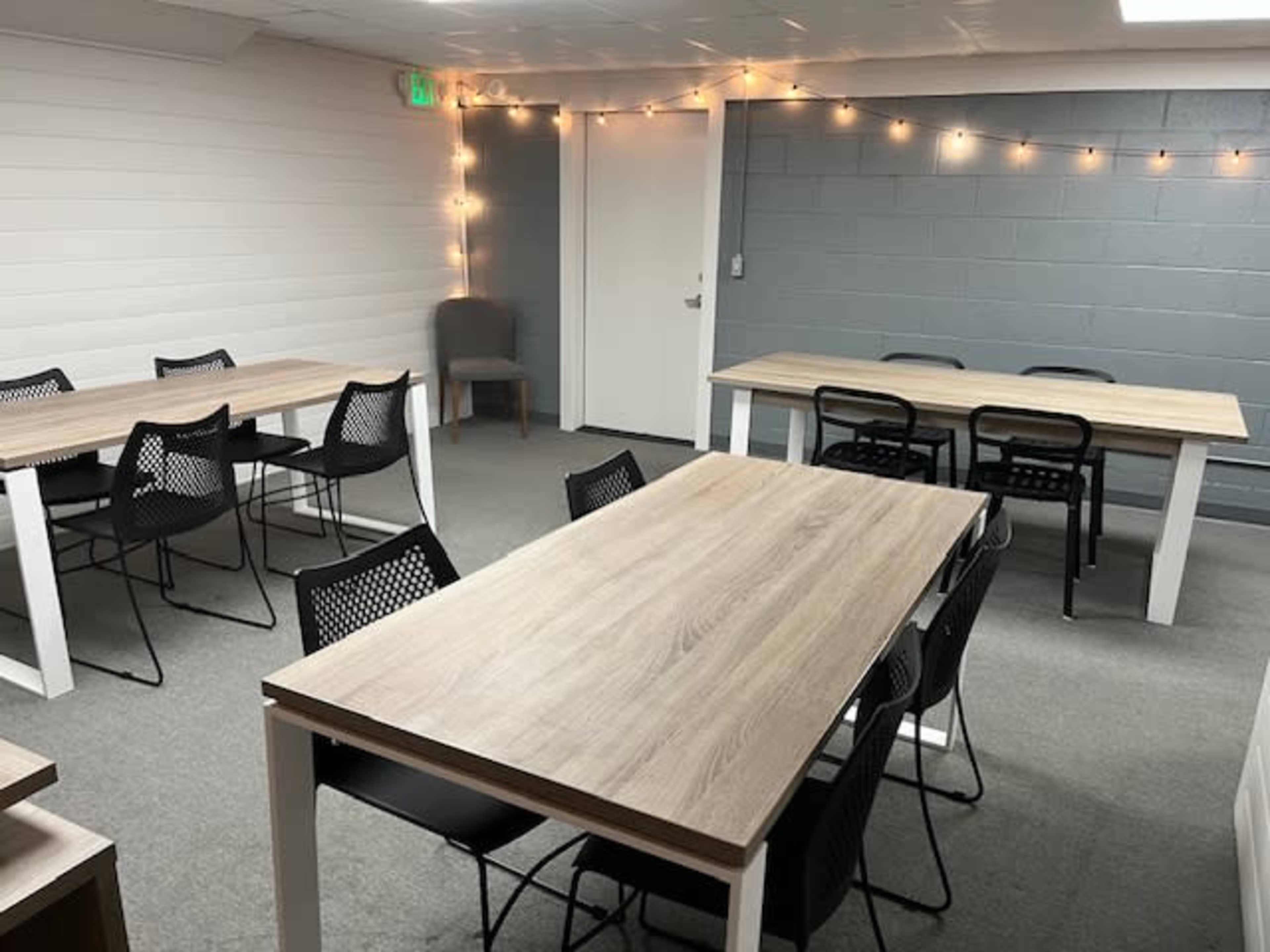 A room with multiple wooden tables and black chairs, illuminated by string lights hanging on the walls.