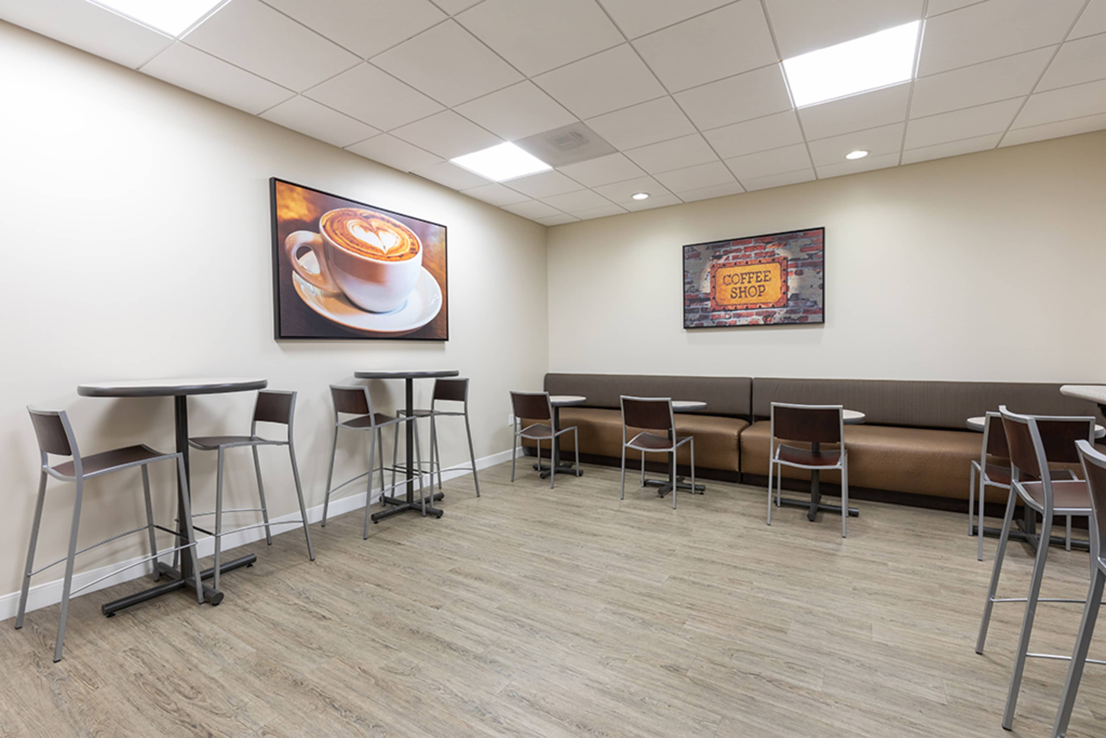 The image shows a small coffee shop area with several high tables and chairs, a brown bench along one wall, and two coffee-themed artworks on the walls.