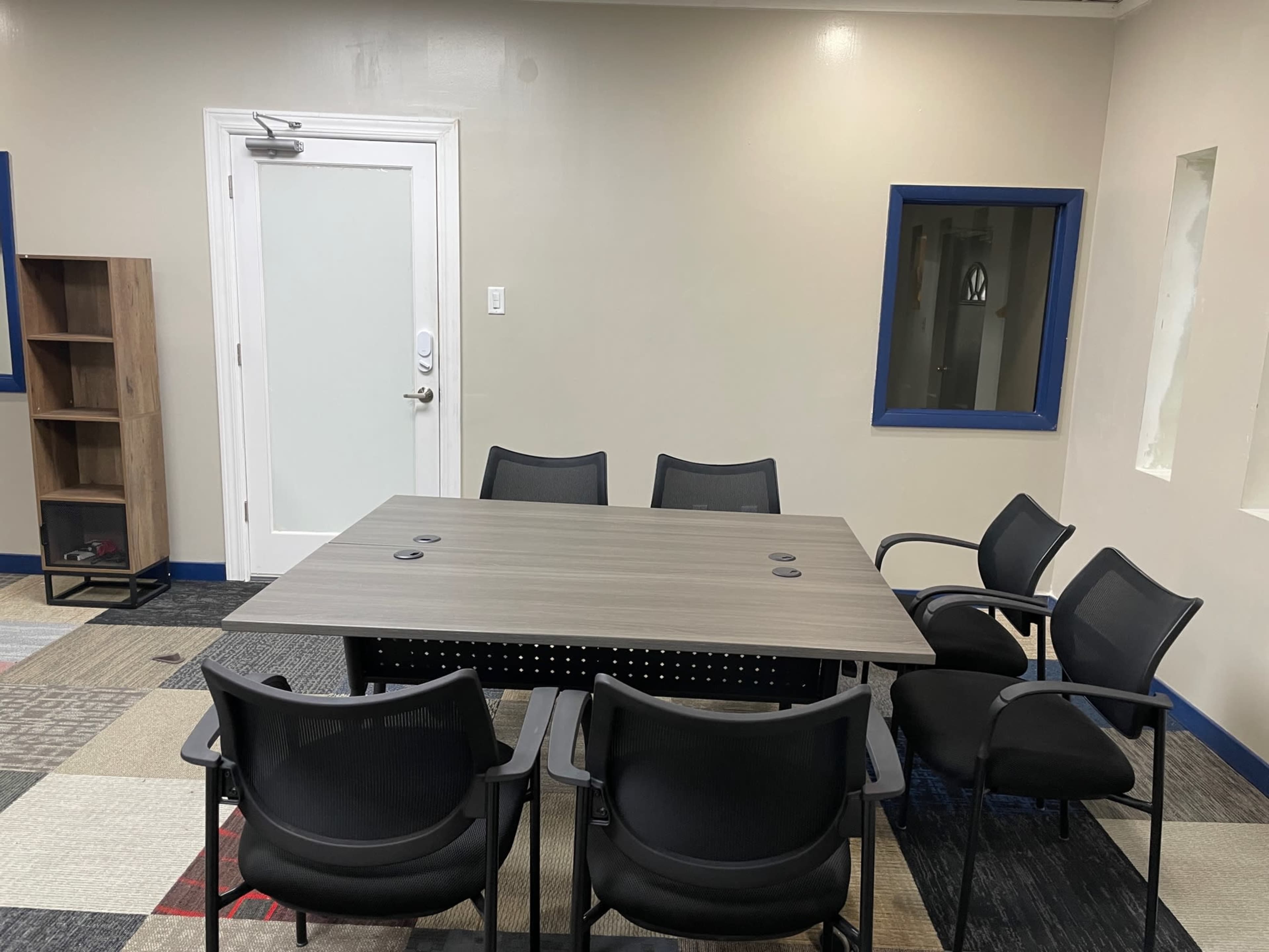 A conference room features a large table surrounded by six black chairs, with a door and two windows on the walls.