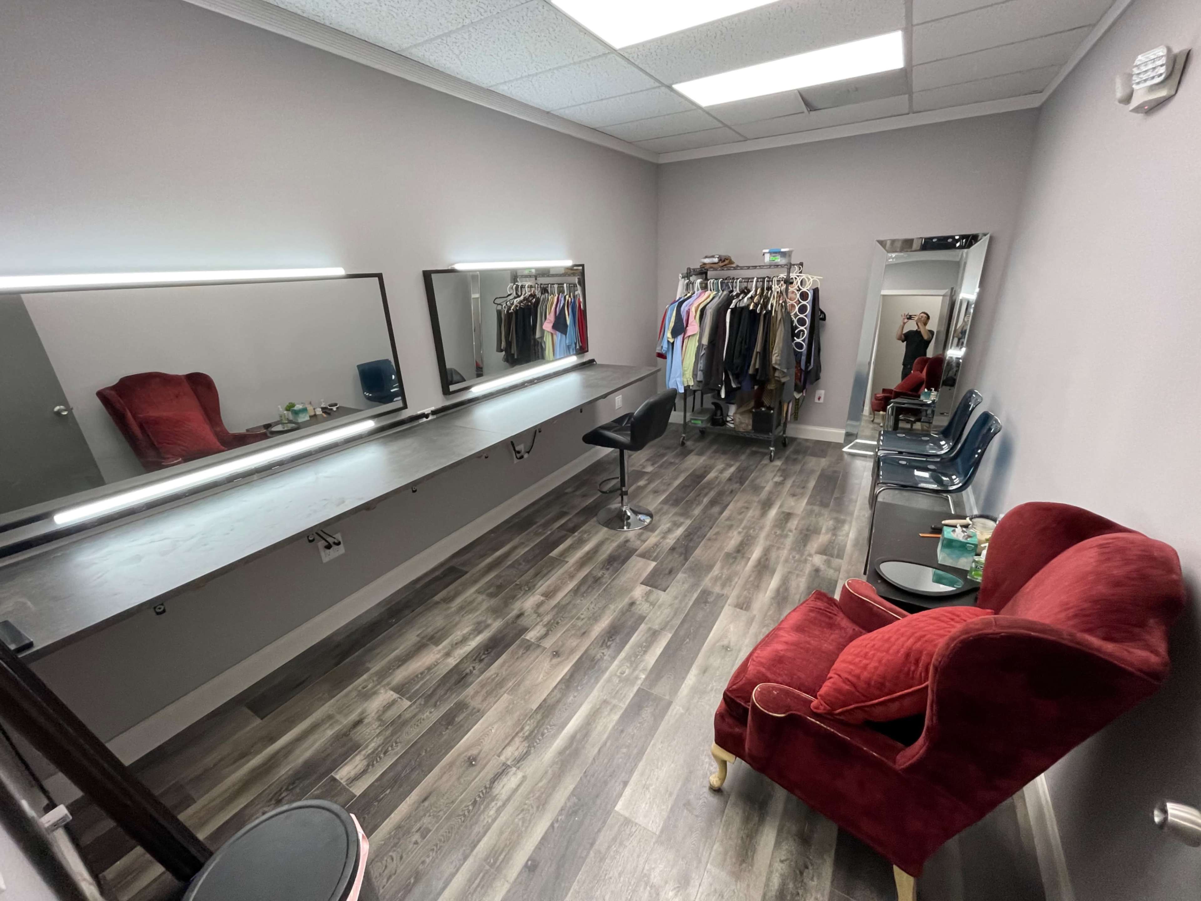 A dressing room with a long mirror, a stool, a red chair, and a rack of hanging clothes against a gray wall.