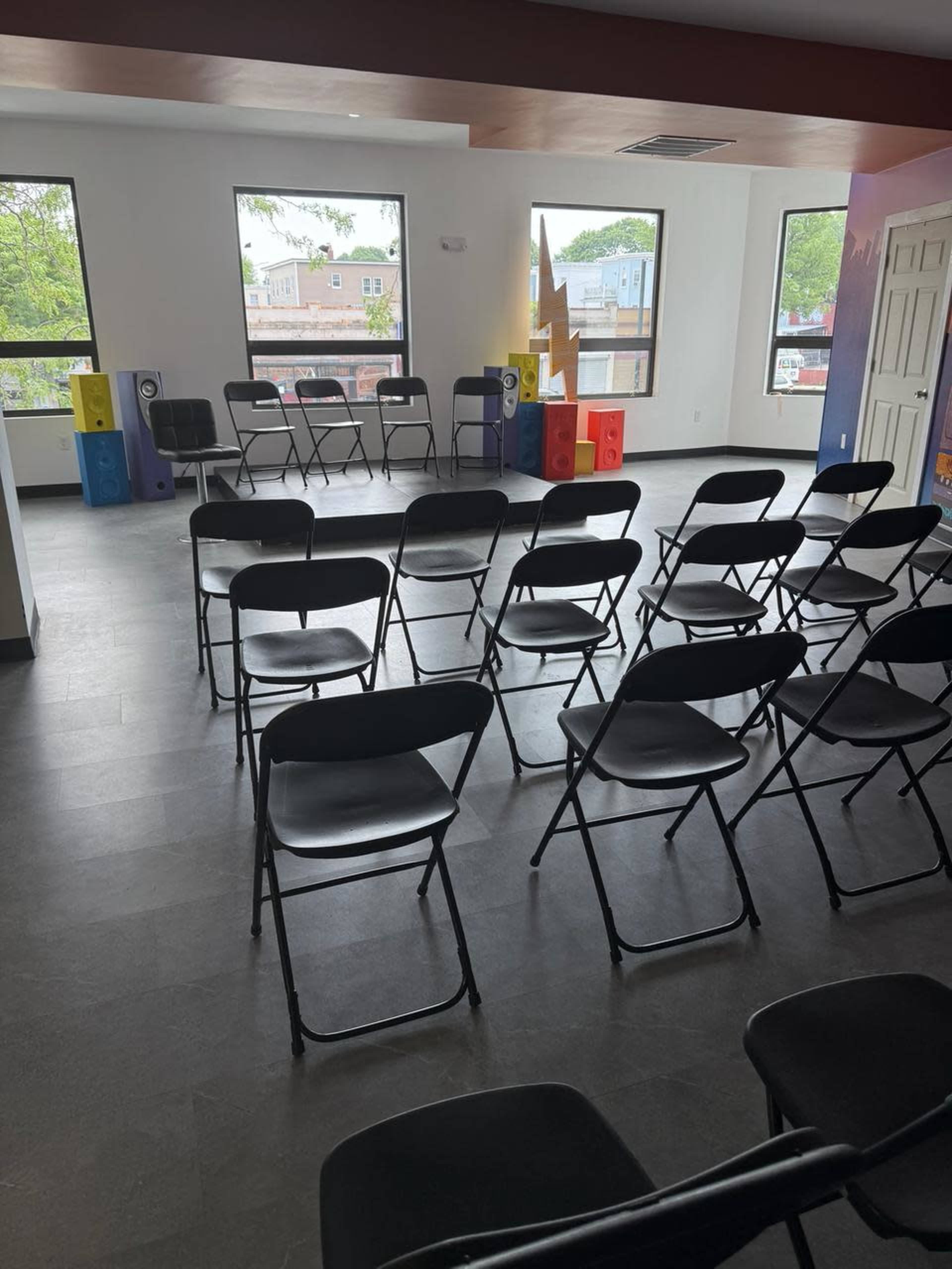 A room with several rows of black folding chairs facing a simple table, decorated with colorful speakers, large windows, and a mural on the wall.
