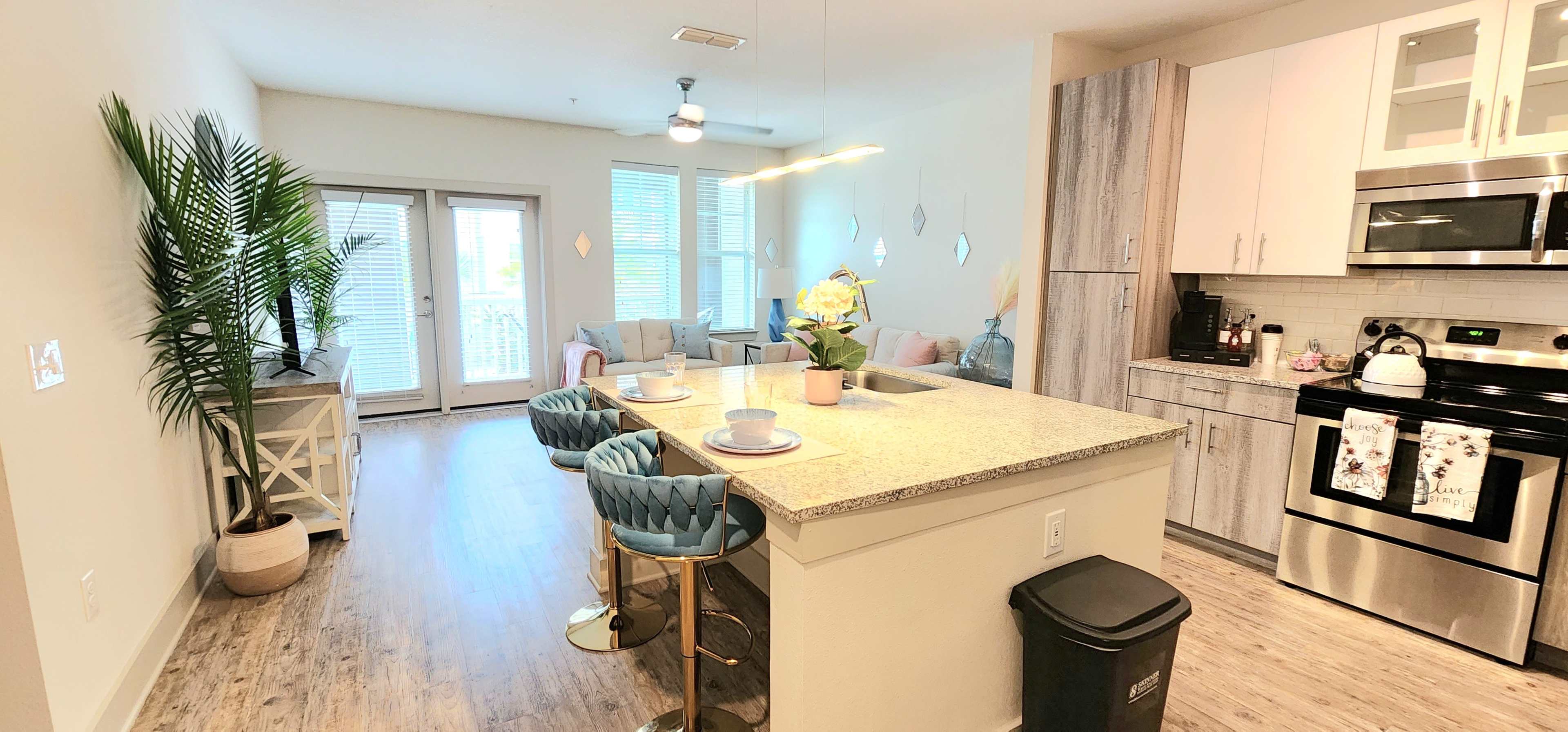 A modern kitchen and dining area with a breakfast bar, light-colored cabinetry, and a living room beyond featuring large windows and comfortable seating.
