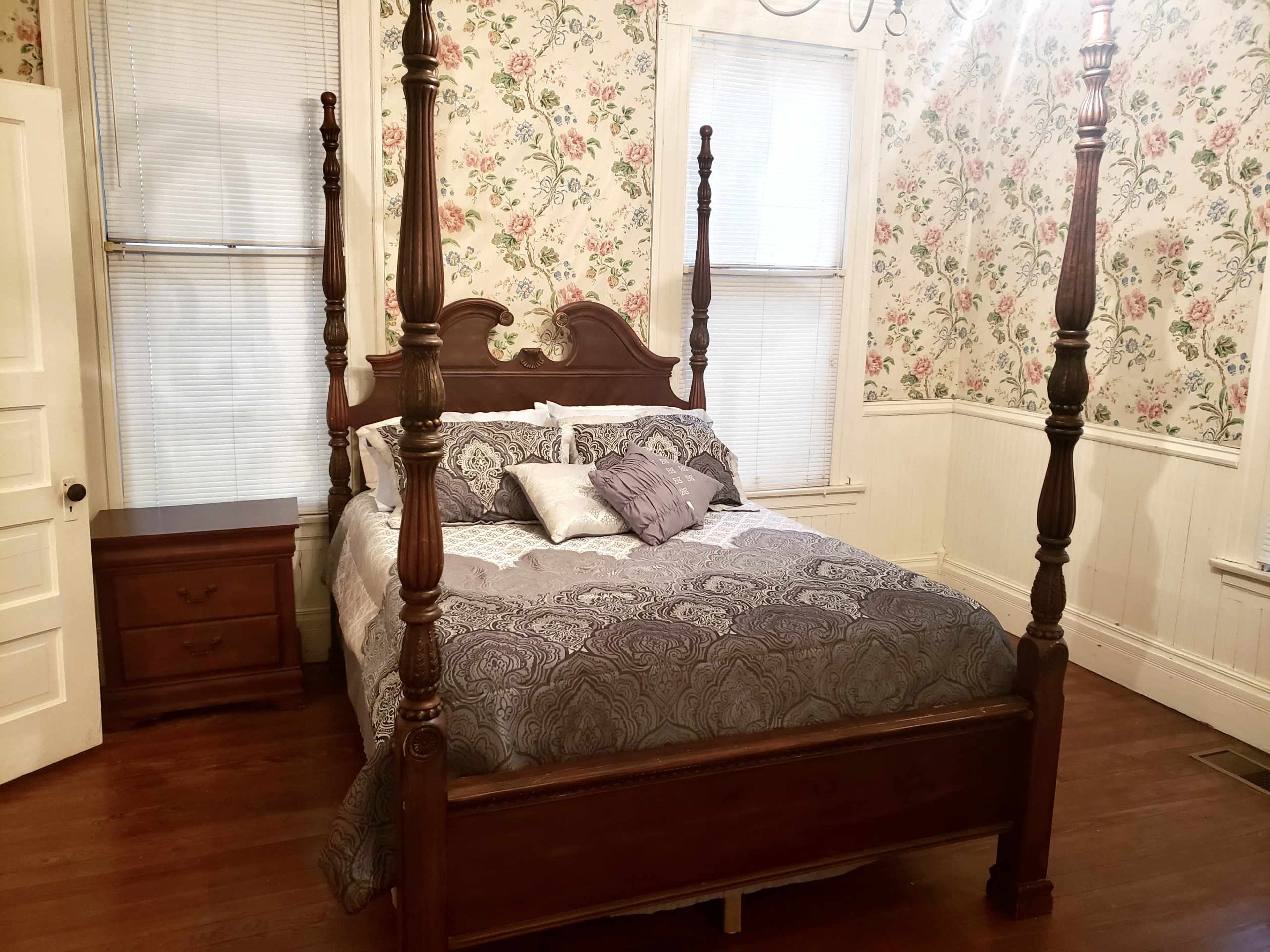 The image shows a bedroom with a four-poster wooden bed, a nightstand, and floral wallpaper.