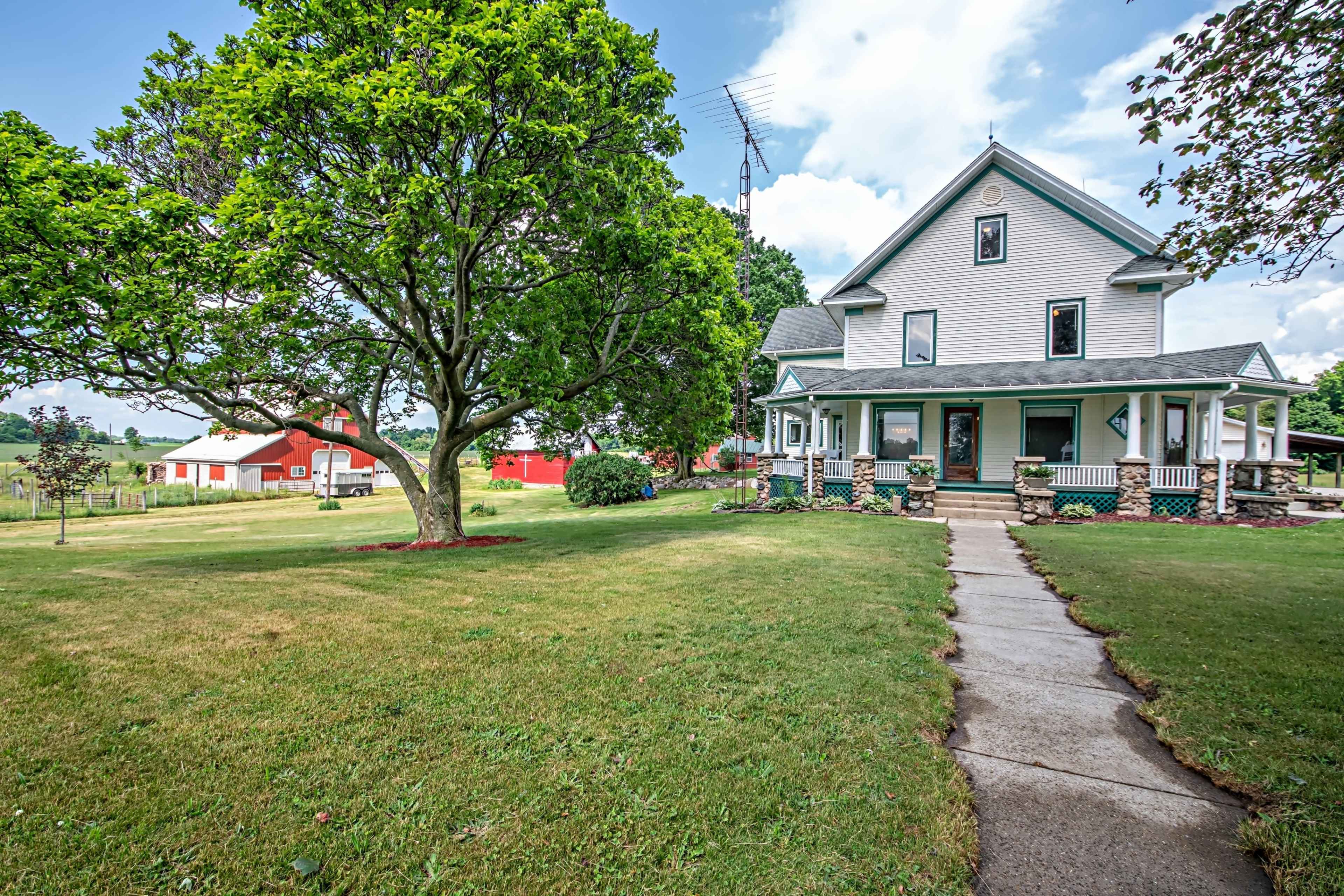 A large, two-story farmhouse with a wraparound porch is situated on a grassy lawn, accompanied by trees and a red barn in the background.