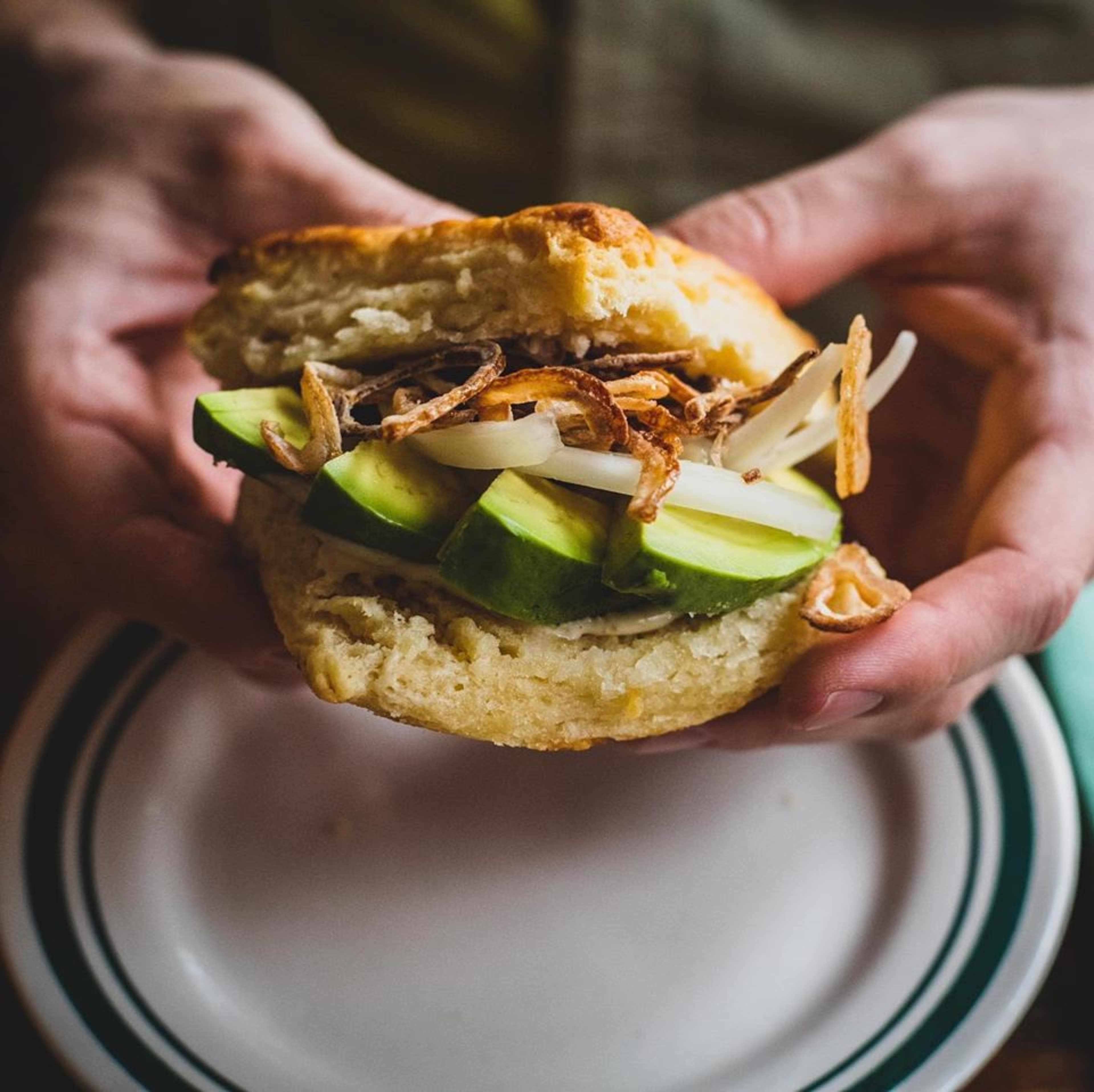 A person holds a biscuit sandwich filled with slices of cucumber and crispy onions.