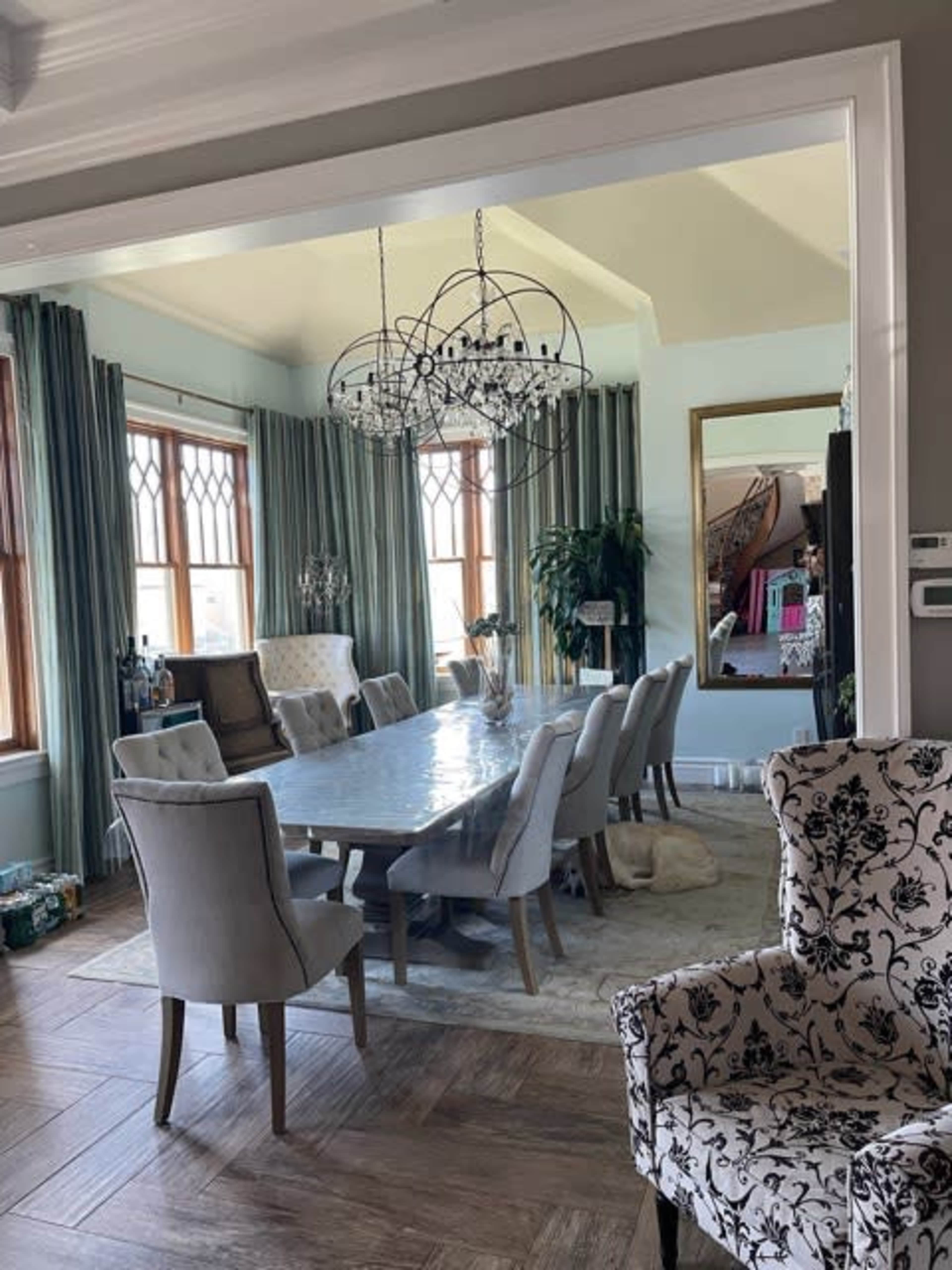 A spacious dining room features a large table surrounded by upholstered chairs, a chandelier overhead, and large windows with green drapery.