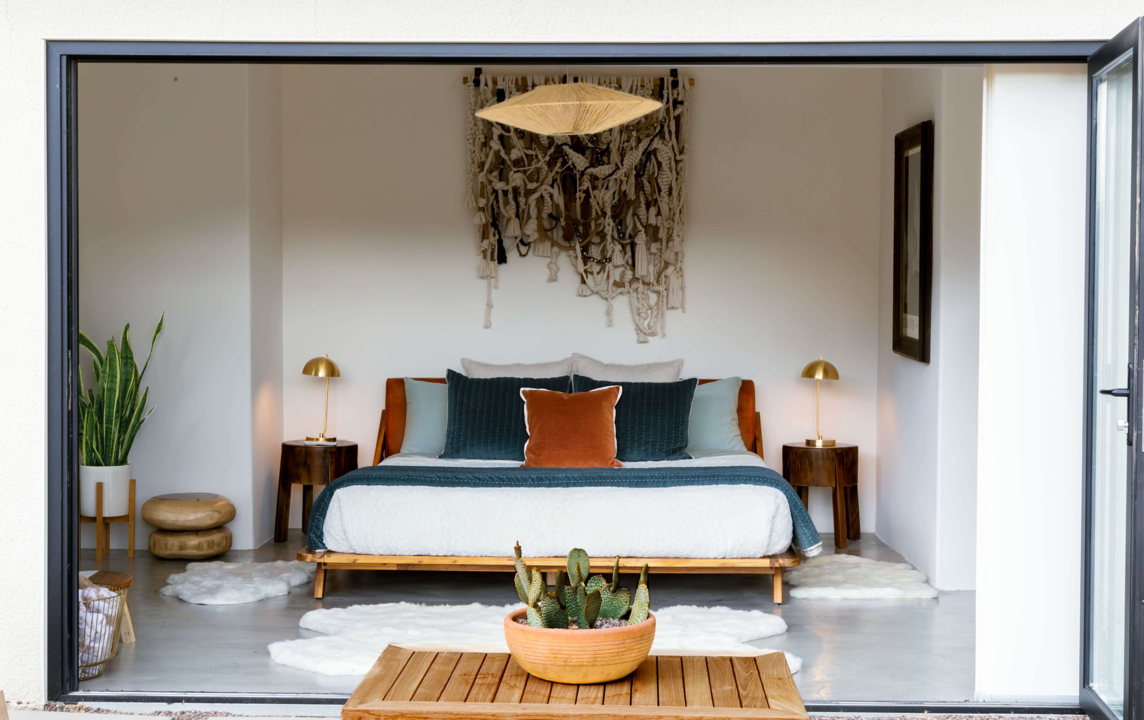 A modern bedroom features a large bed with a blue and white duvet, flanked by two bedside lamps, with a decorative macramé wall hanging in the background and a cactus centerpiece on a wooden table.