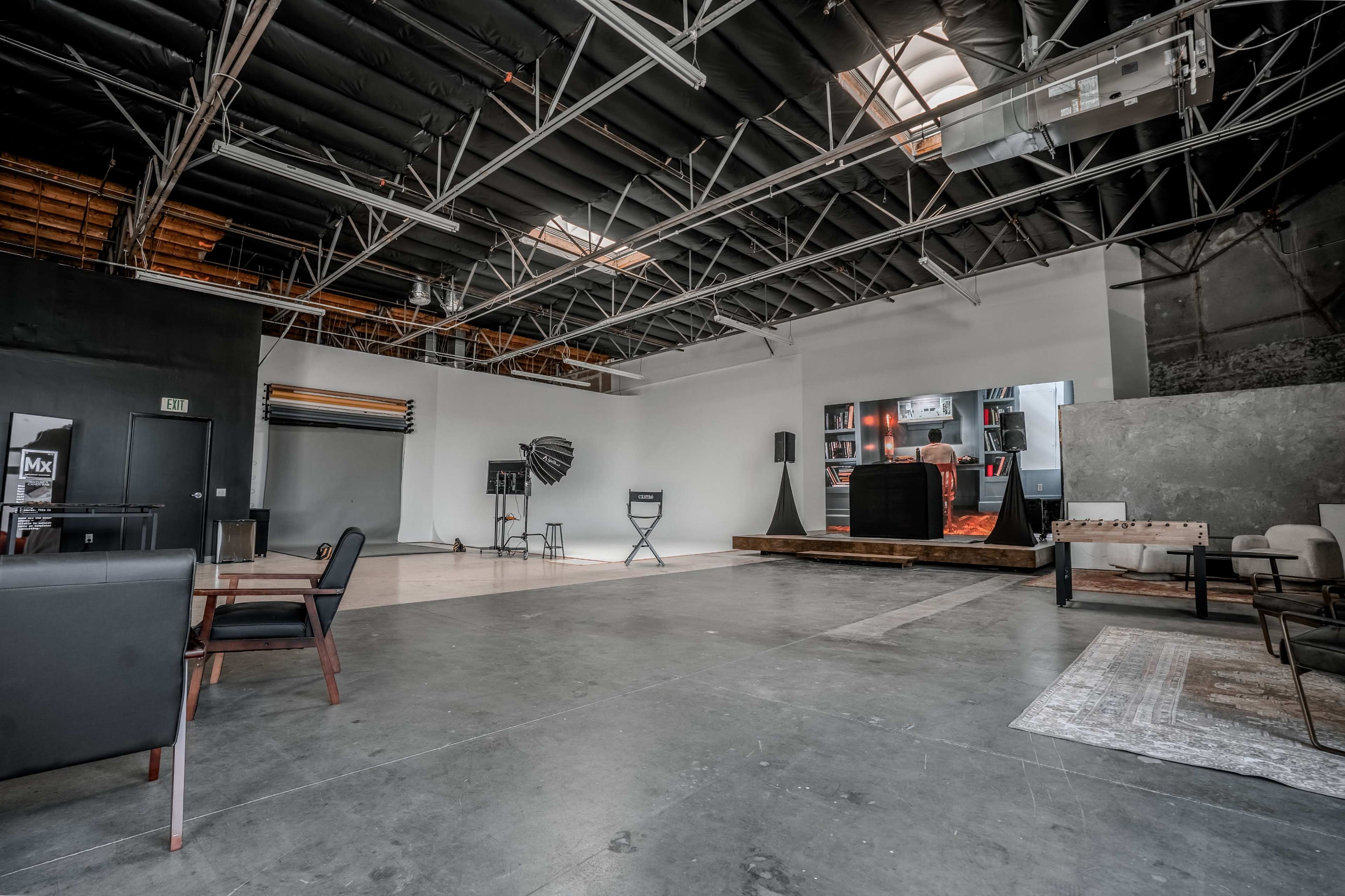 The image shows a spacious, industrial-style room with high ceilings and concrete floors, featuring minimal furniture and a stage area at one end.