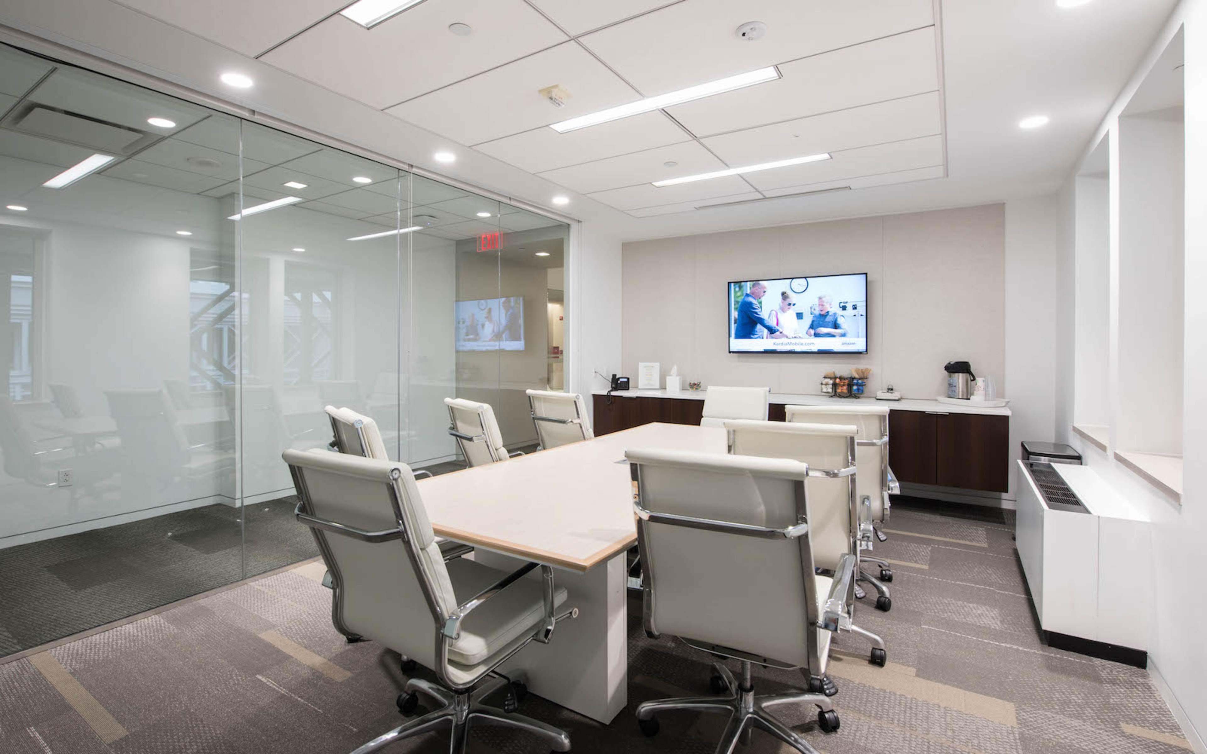 A modern conference room features a central table surrounded by white ergonomic chairs, glass walls, and a wall-mounted television displaying a presentation.