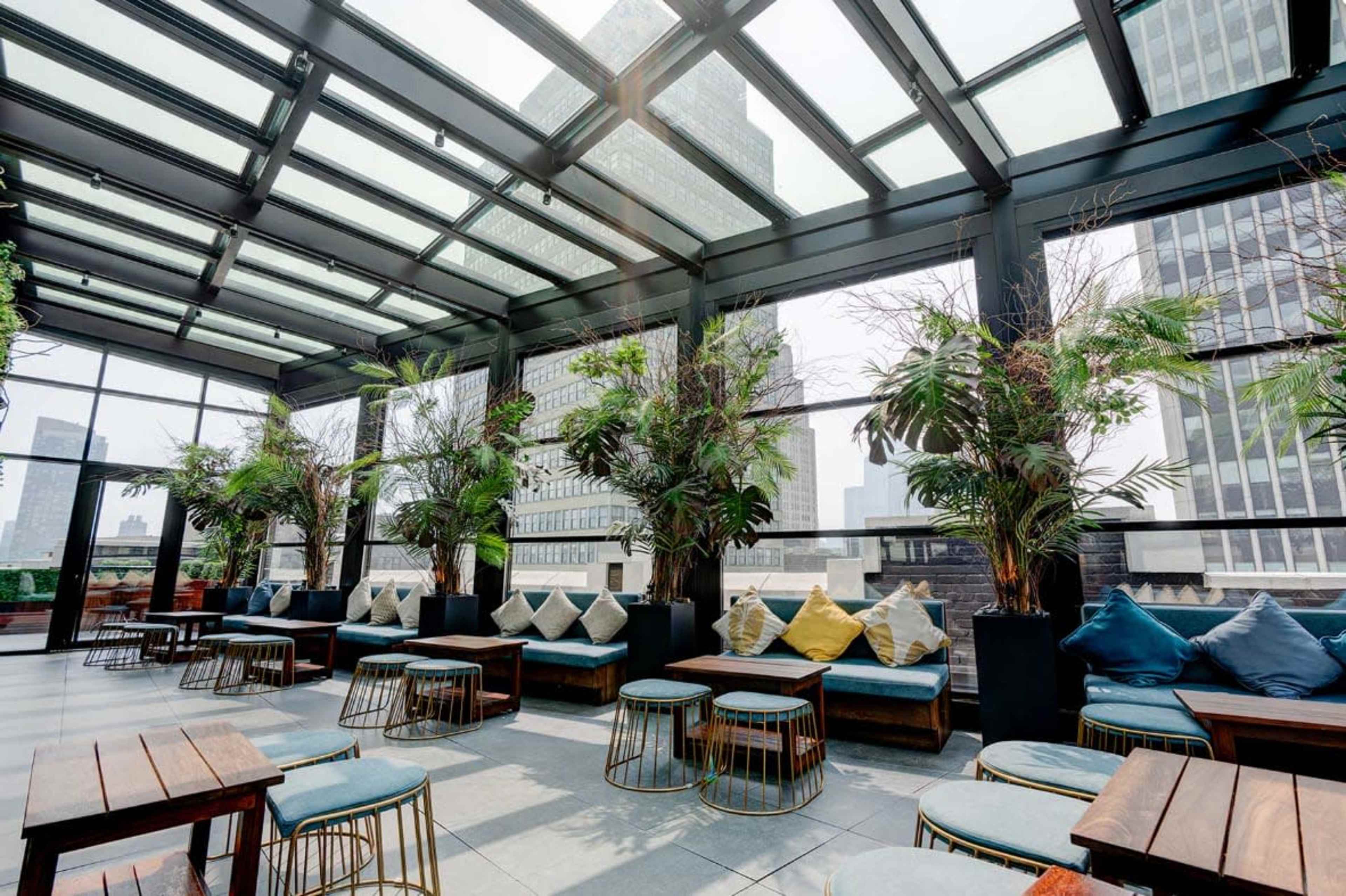 The image shows a modern rooftop lounge with glass walls, featuring plants, comfortable seating, and tables, overlooking a cityscape.