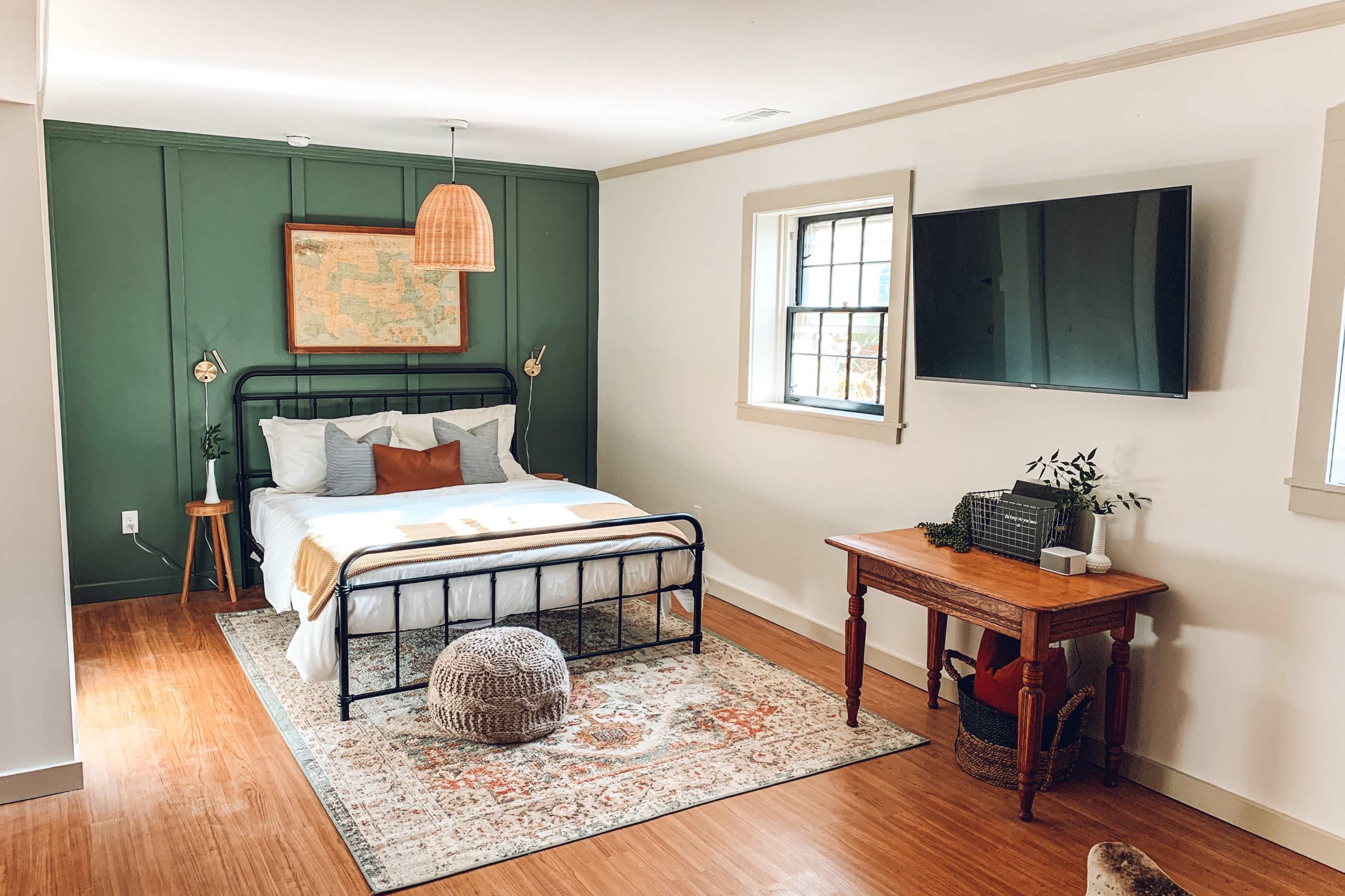 A cozy bedroom features a black metal bed frame with white bedding, a green accent wall, a decorative rug, and a wooden table next to a television.