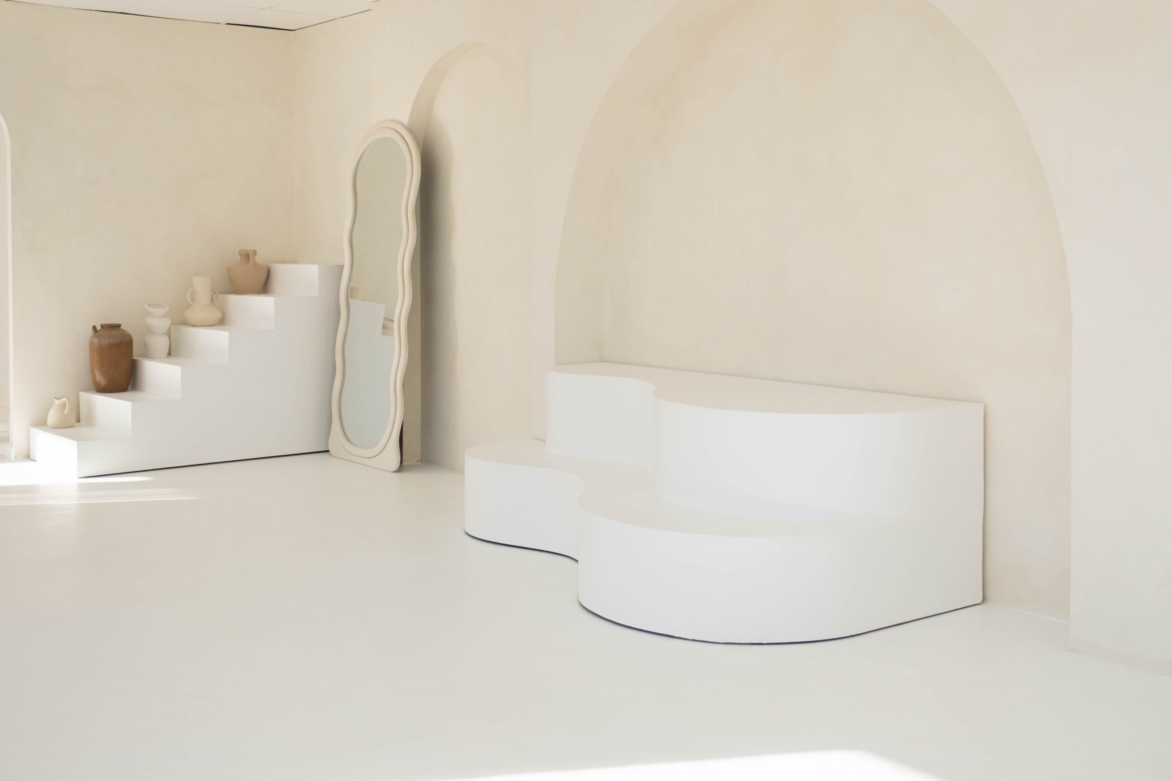 The scene features a minimalist interior with curved white furniture, a decorative mirror, and steps leading to a ledge displaying various pottery pieces.