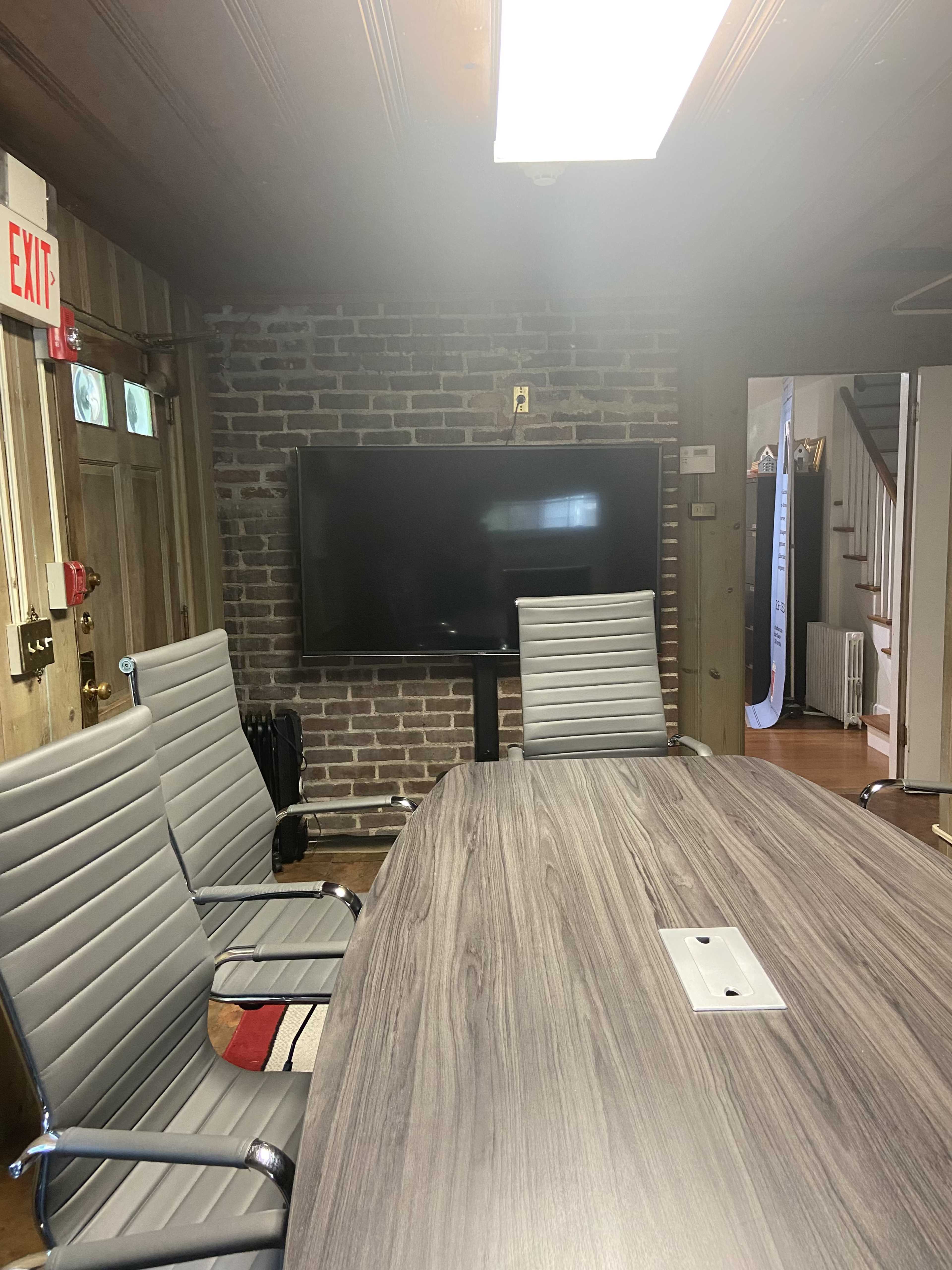 A conference room features a long table surrounded by sleek chairs, with a large flat-screen TV mounted on a brick wall.