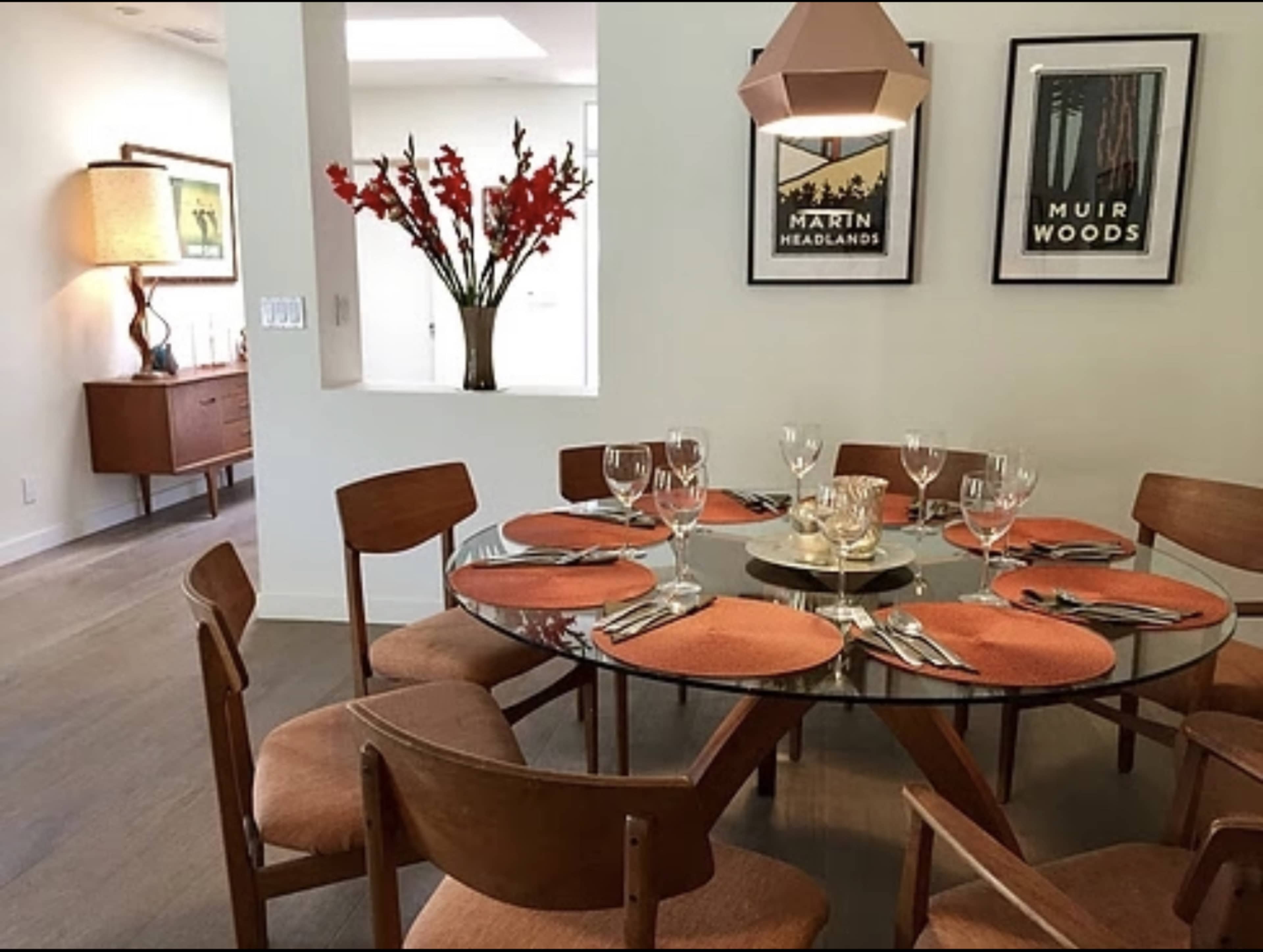 A modern dining room featuring a round table set with orange placemats and glasses, surrounded by wooden chairs, with framed artworks on the wall and a decorative lamp in the background.