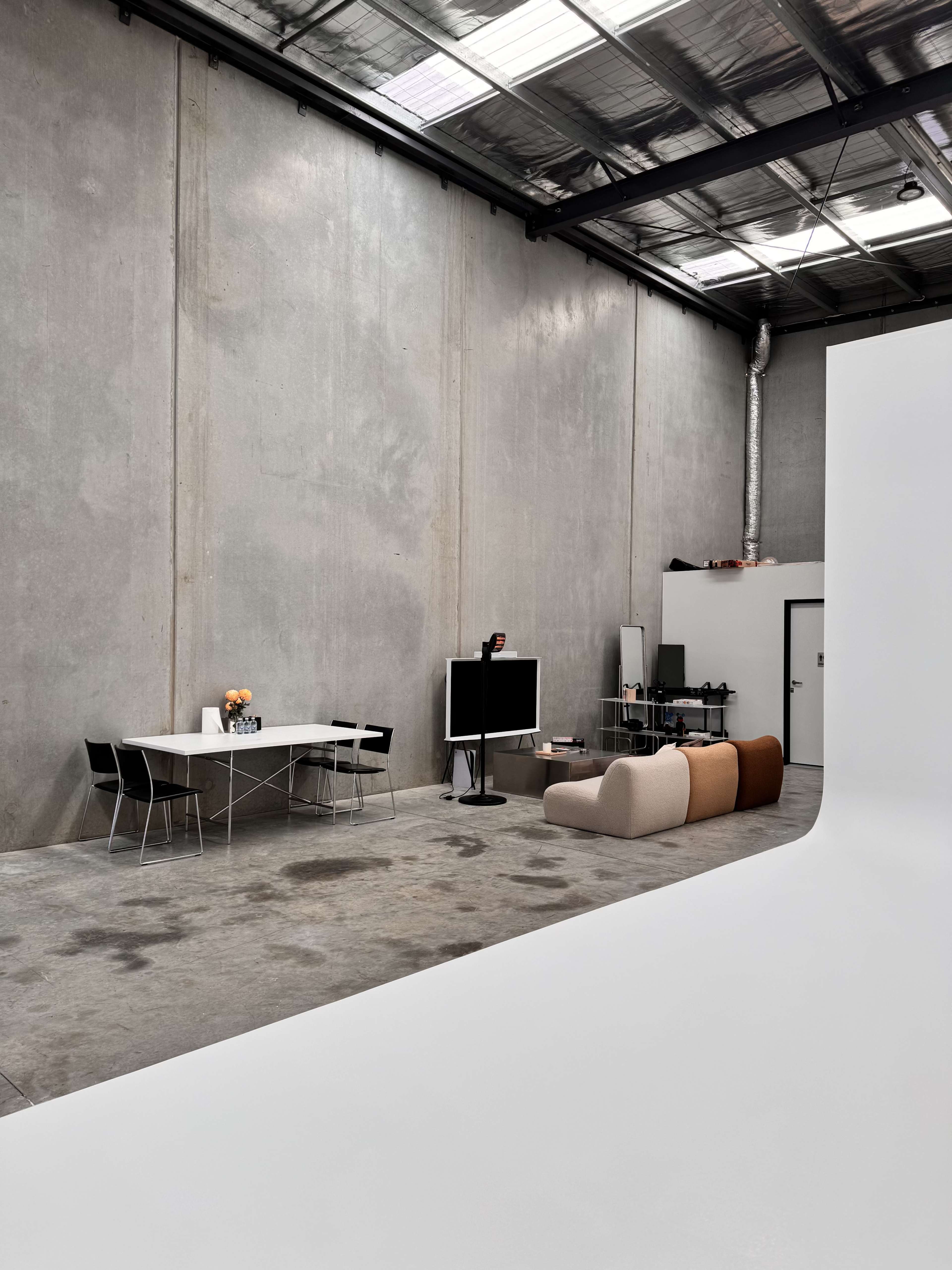 The image shows an industrial-style interior with a concrete wall, a dining table with chairs, a television, and a curved sofa in a spacious room.
