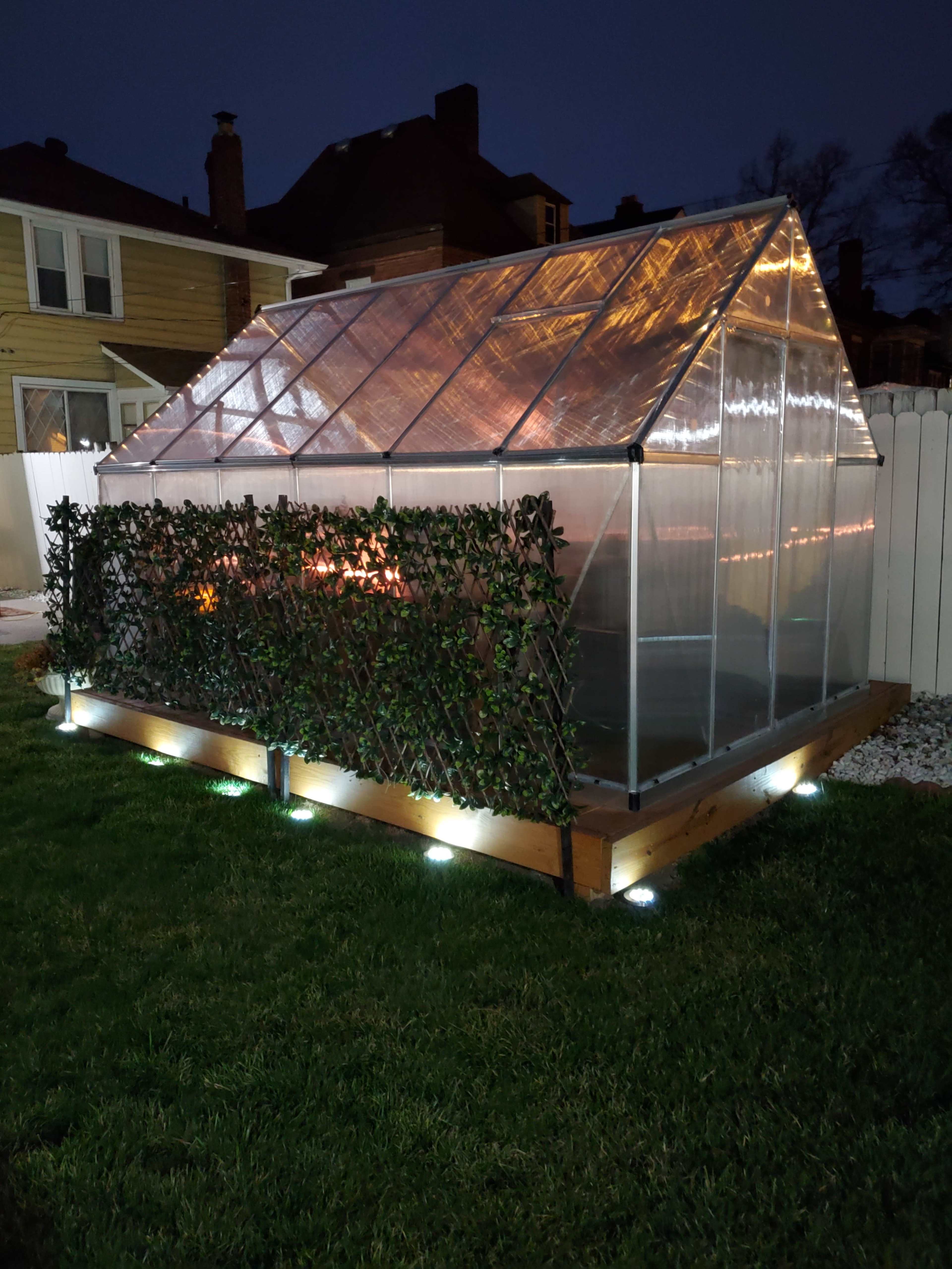 A greenhouse with transparent panels and a wooden base is illuminated at night, surrounded by a green hedge and positioned on a lawn.