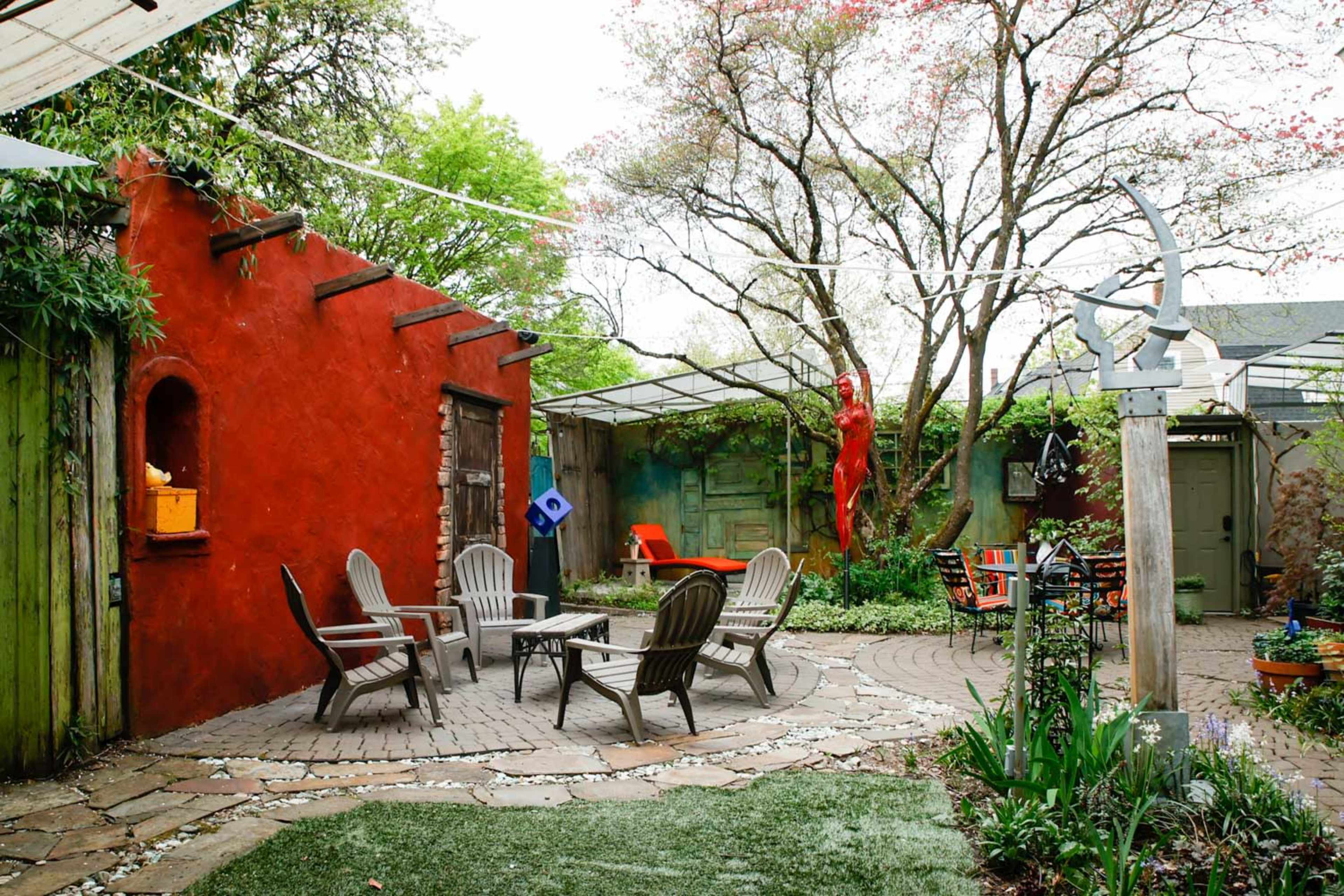 An outdoor garden space featuring a circular stone patio with four Adirondack chairs, surrounded by colorful walls, greenery, and a sculpture.