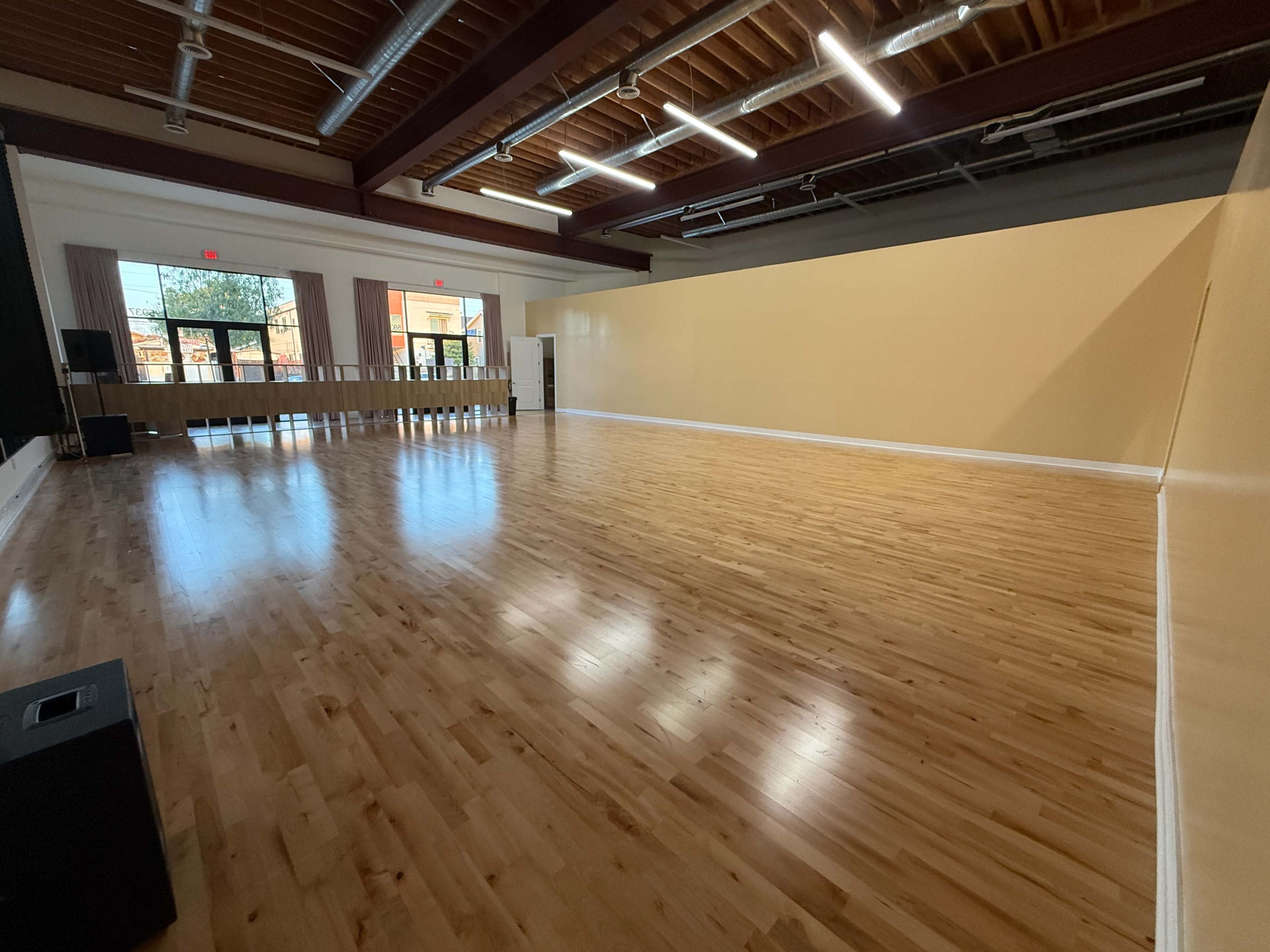 The image shows a spacious, empty studio with wooden flooring, large windows, and a plain yellow wall.