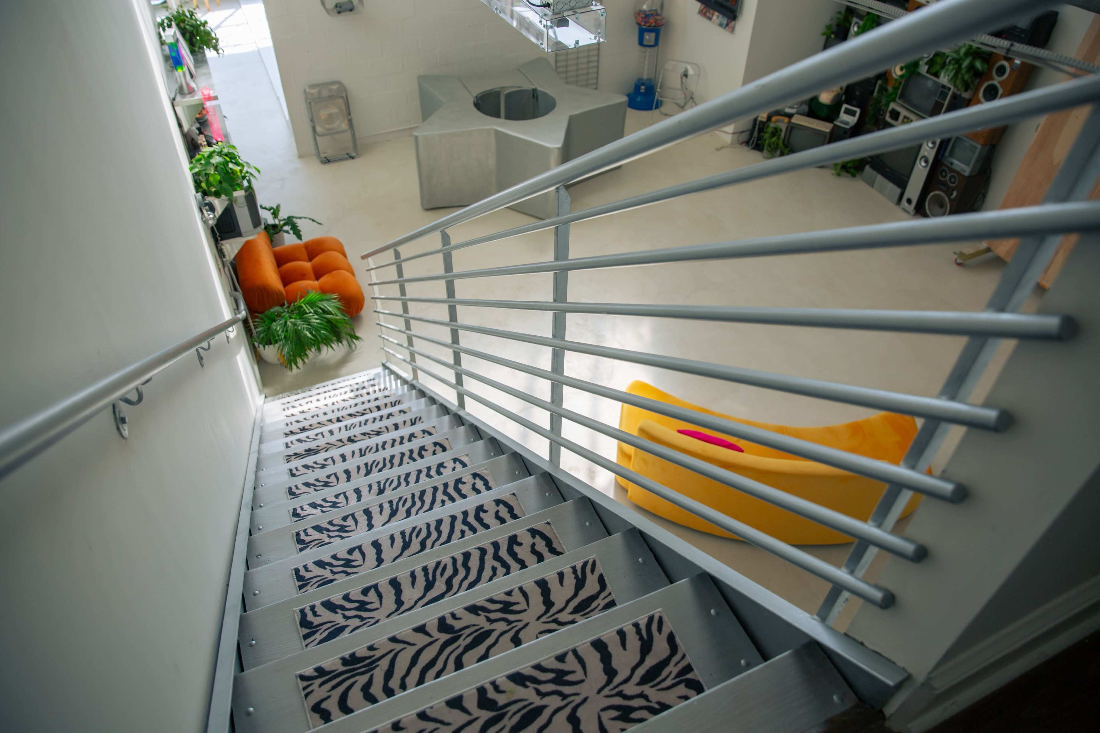 The image shows a staircase with a patterned runner, leading down to a spacious room with modern furniture and plants.
