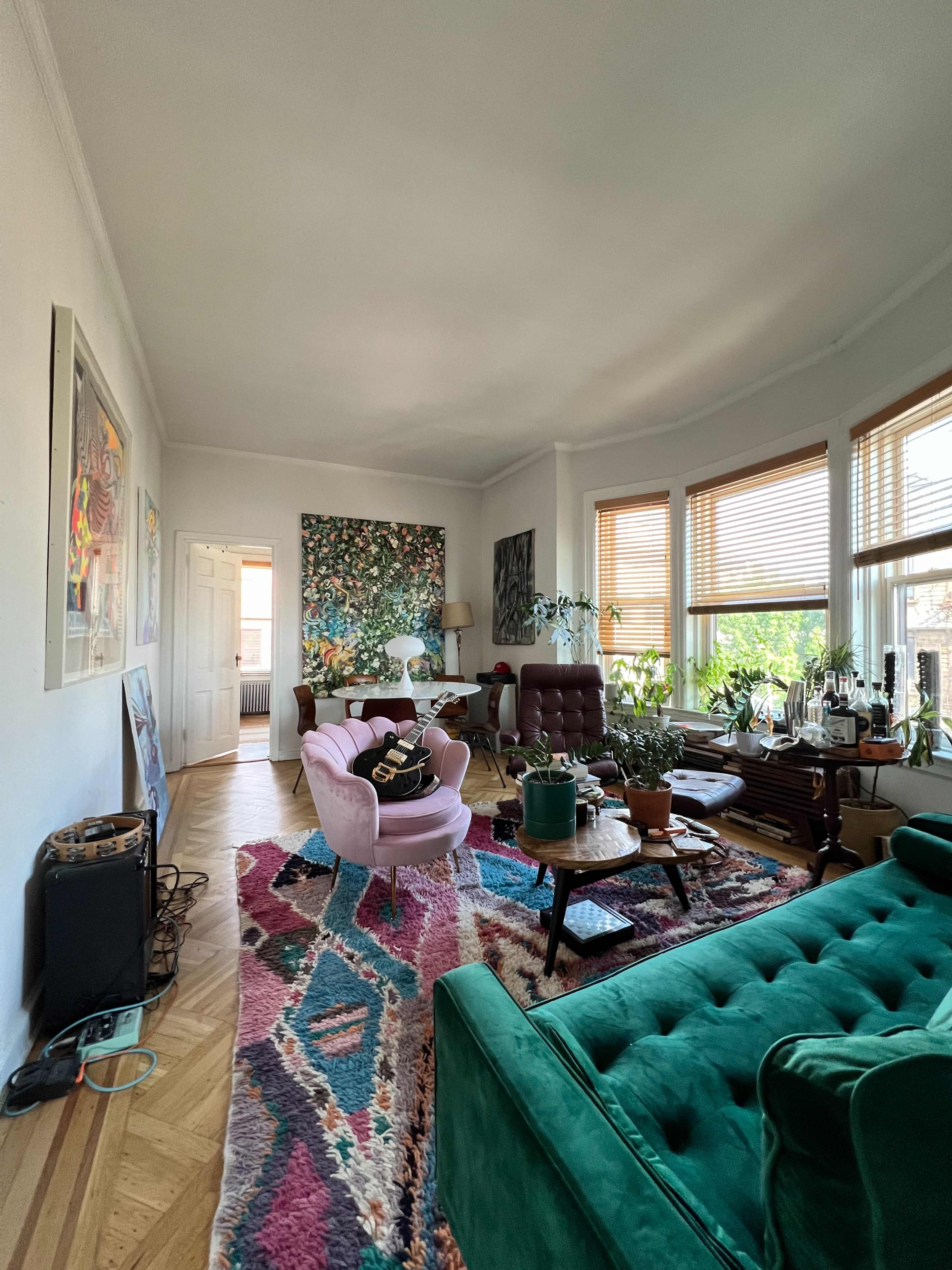 The image shows a bright living room with large windows, a mix of colorful furniture, and a variety of plants.