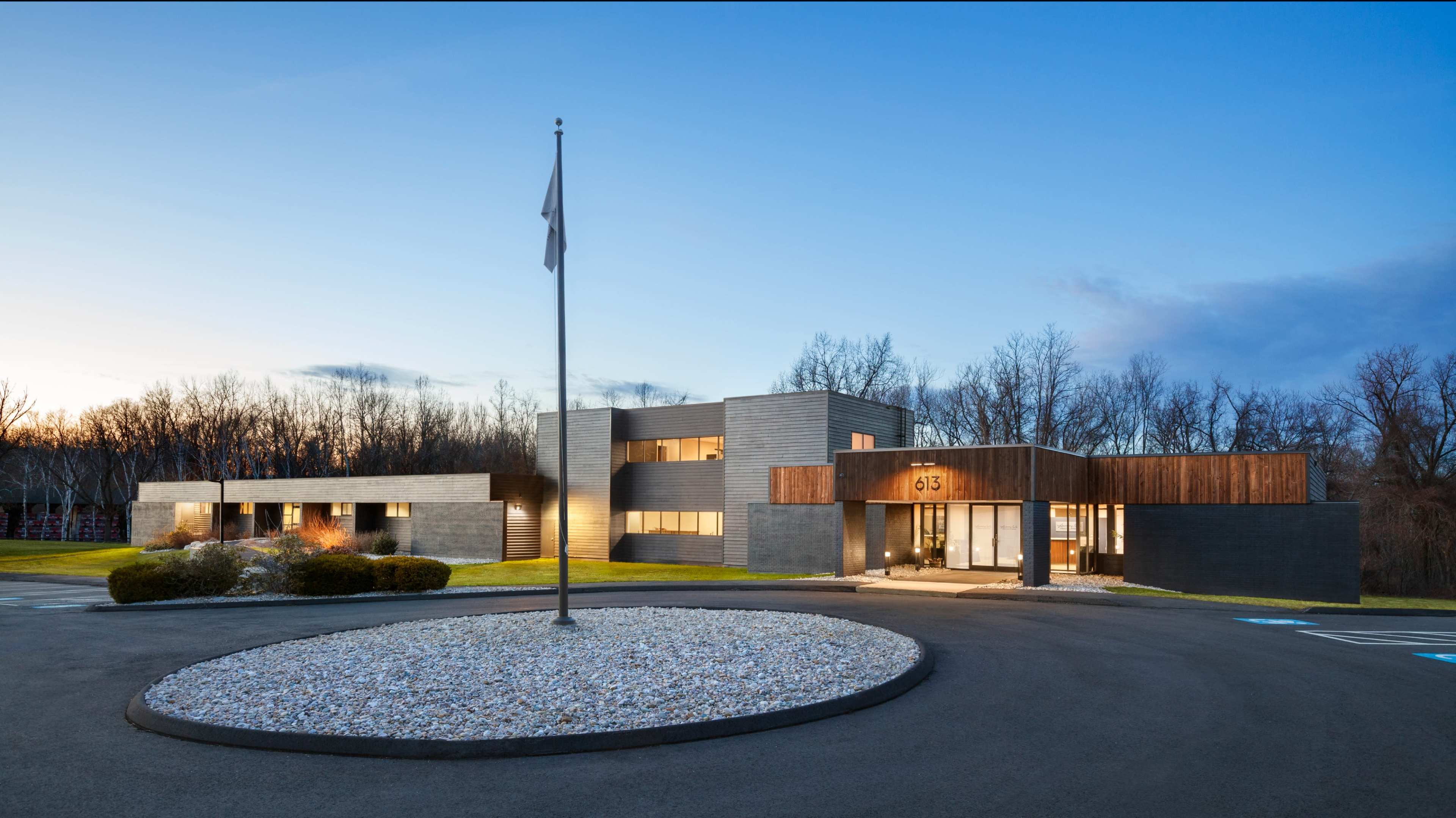 The image shows a modern, multi-level building with a mix of dark and wooden exterior, landscaped grounds, and a circular driveway.