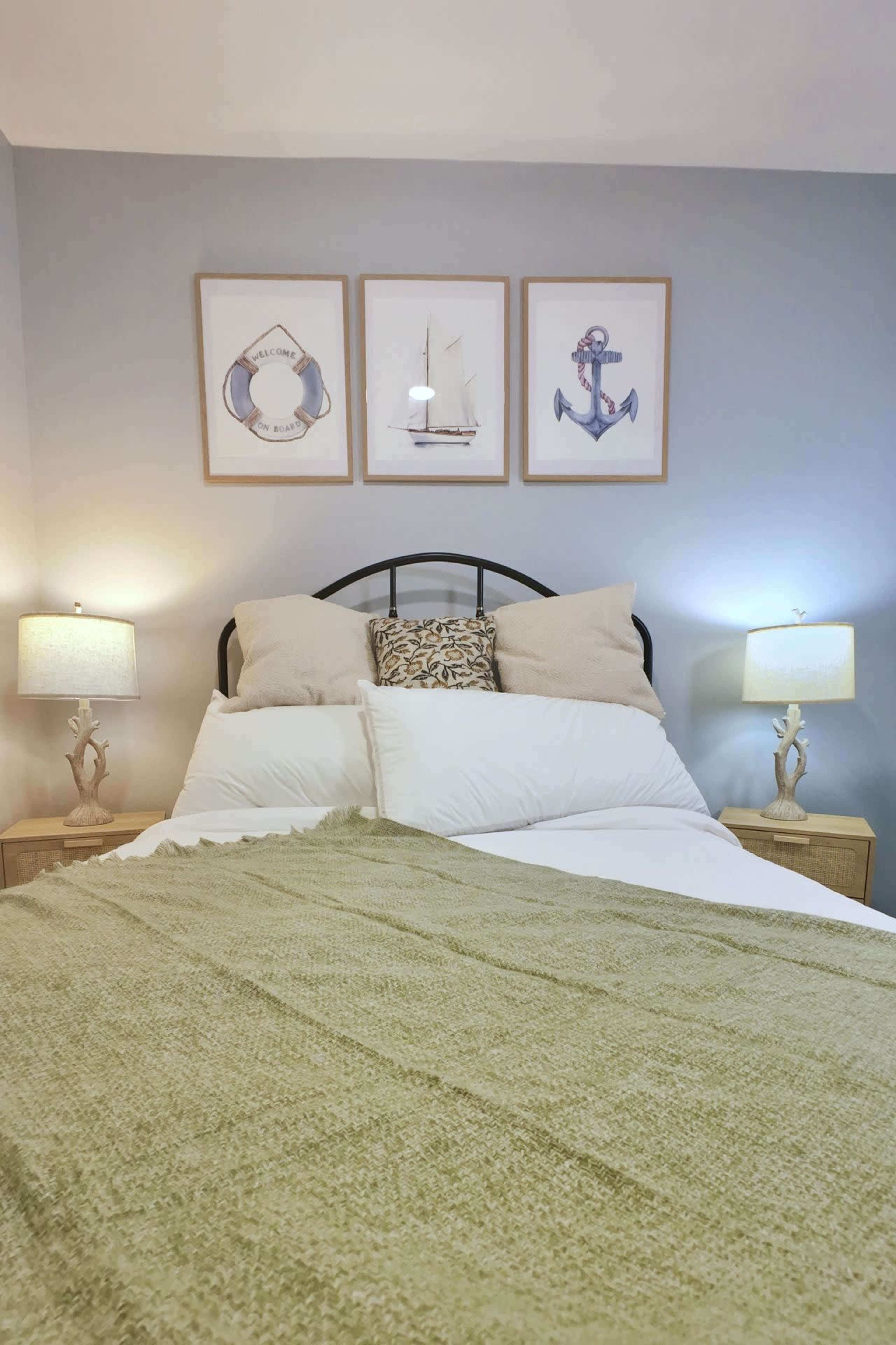 A neatly made bed with a green throw blanket is centered in a room featuring two nightstands with lamps and nautical-themed artwork on the wall.