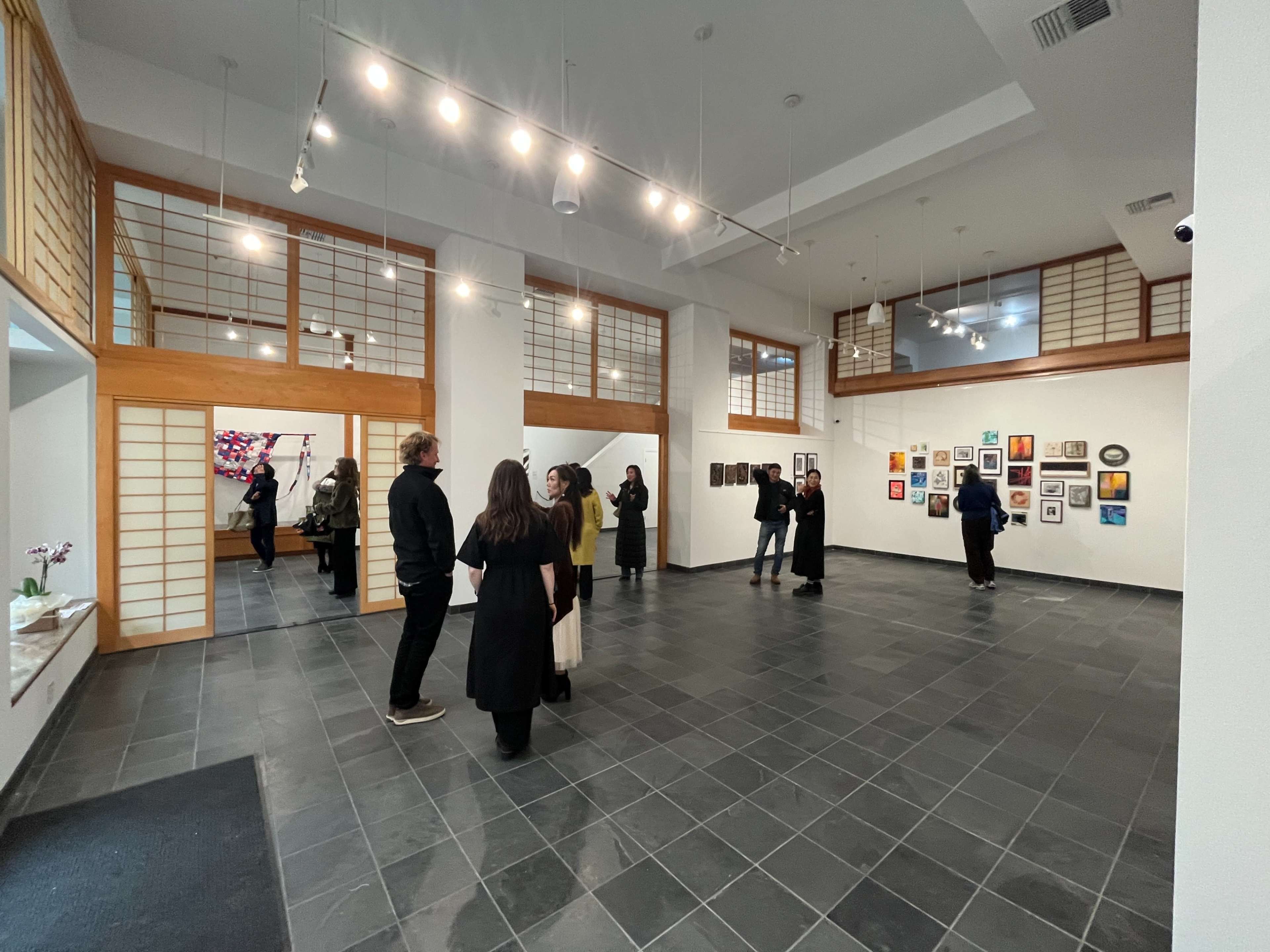 A group of people is observing various artworks displayed on the walls of a spacious, well-lit gallery with a tile floor and wooden accents.