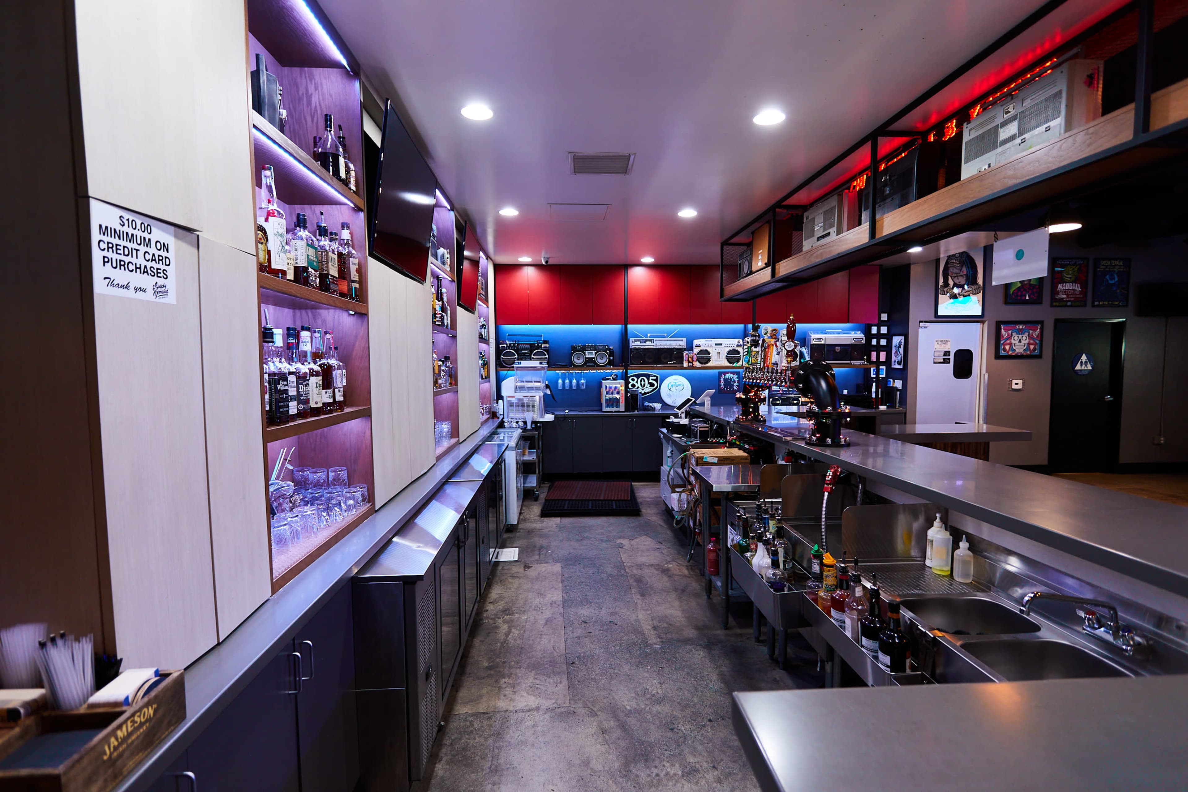 A modern bar area with shelves of liquor bottles, a long counter, and a row of sinks in a well-lit space.