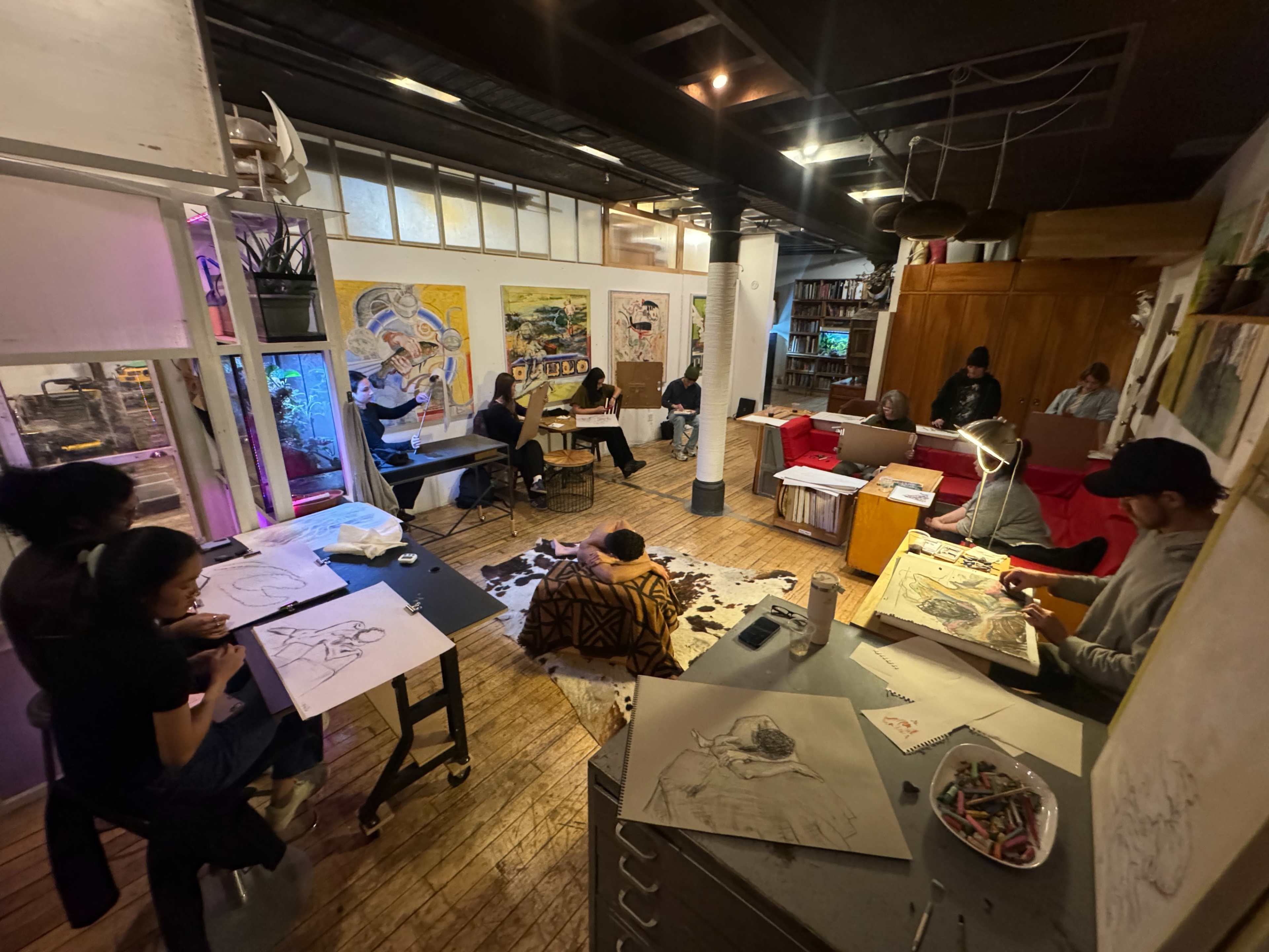 The image shows a creative studio filled with artists working on drawings at various tables in a well-lit environment.