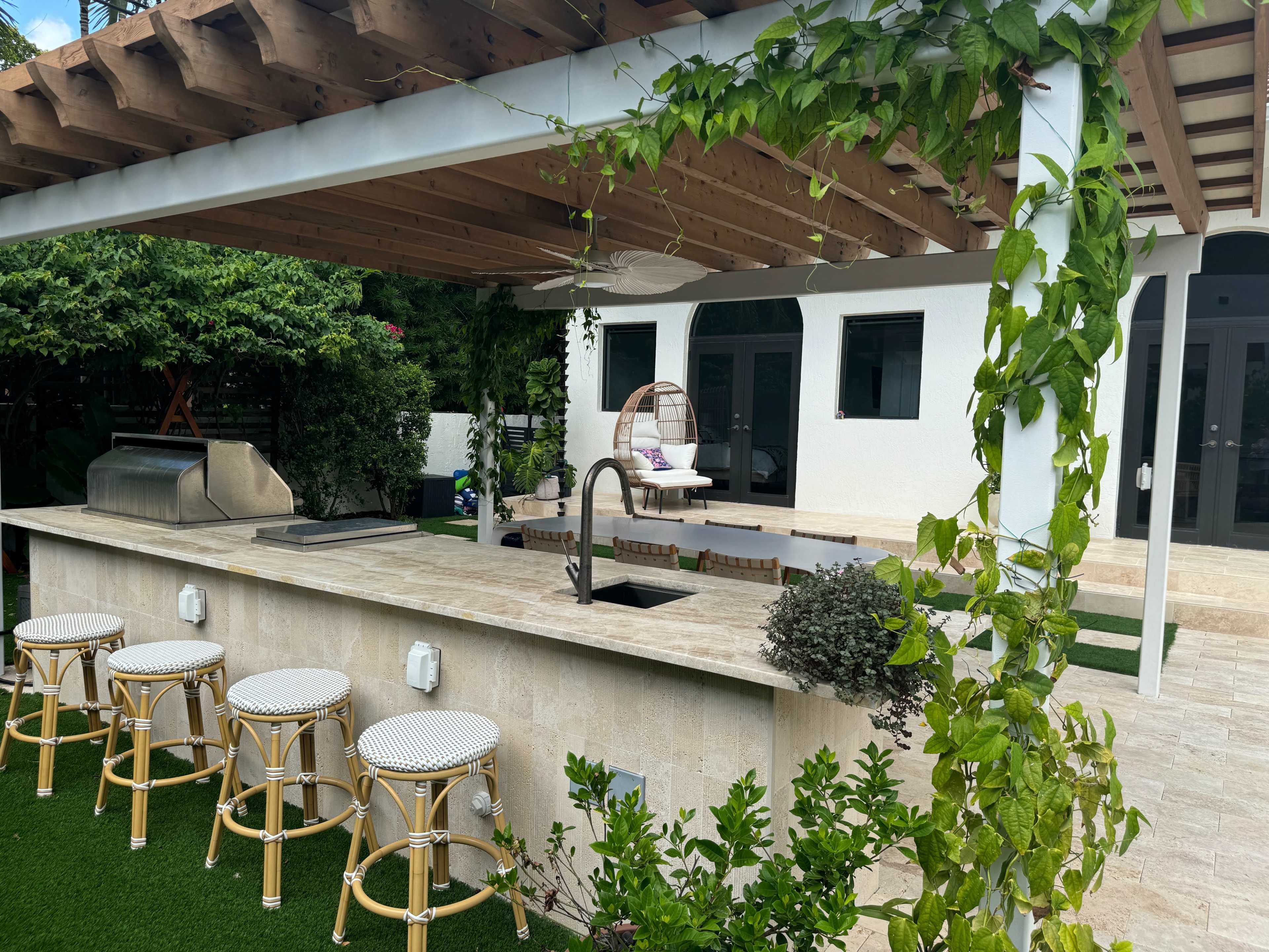 The image shows an outdoor kitchen area with a stone countertop, a grill, and seating surrounded by greenery and a pergola.
