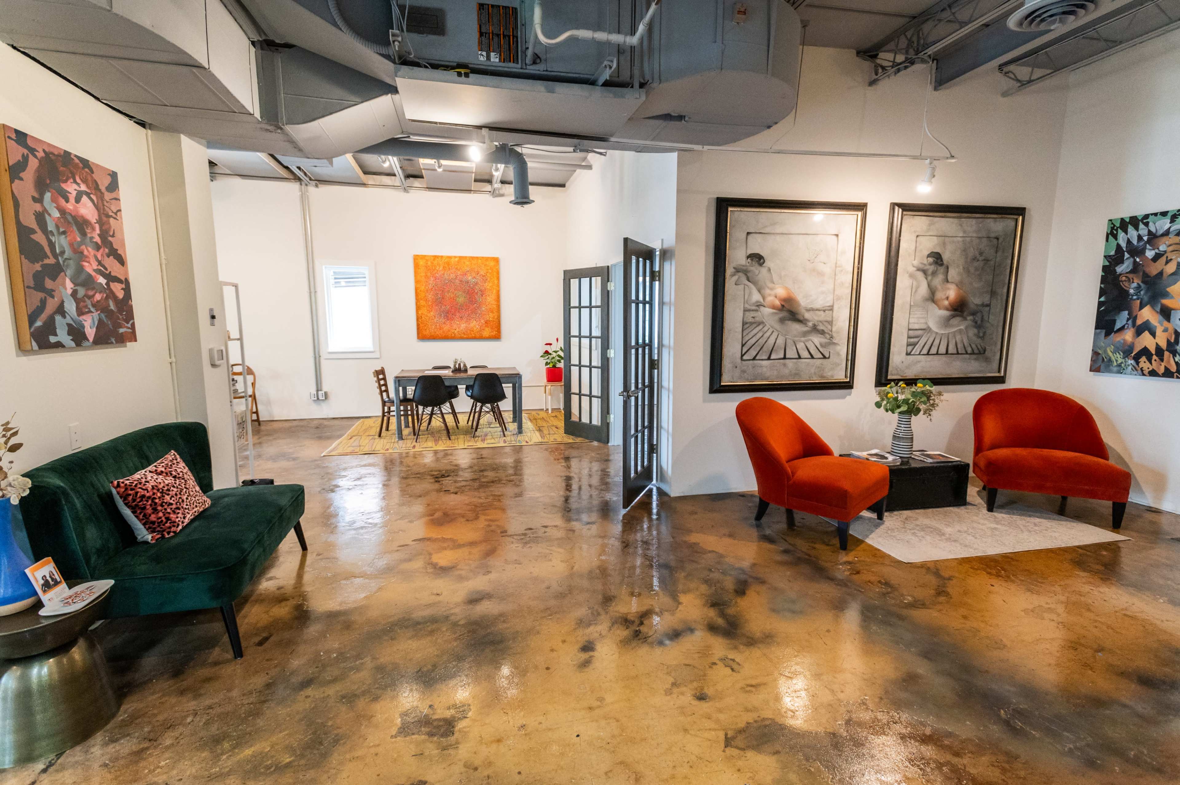 A spacious, modern room with two orange chairs and a green couch, artwork on the walls, and a dining area in the background.