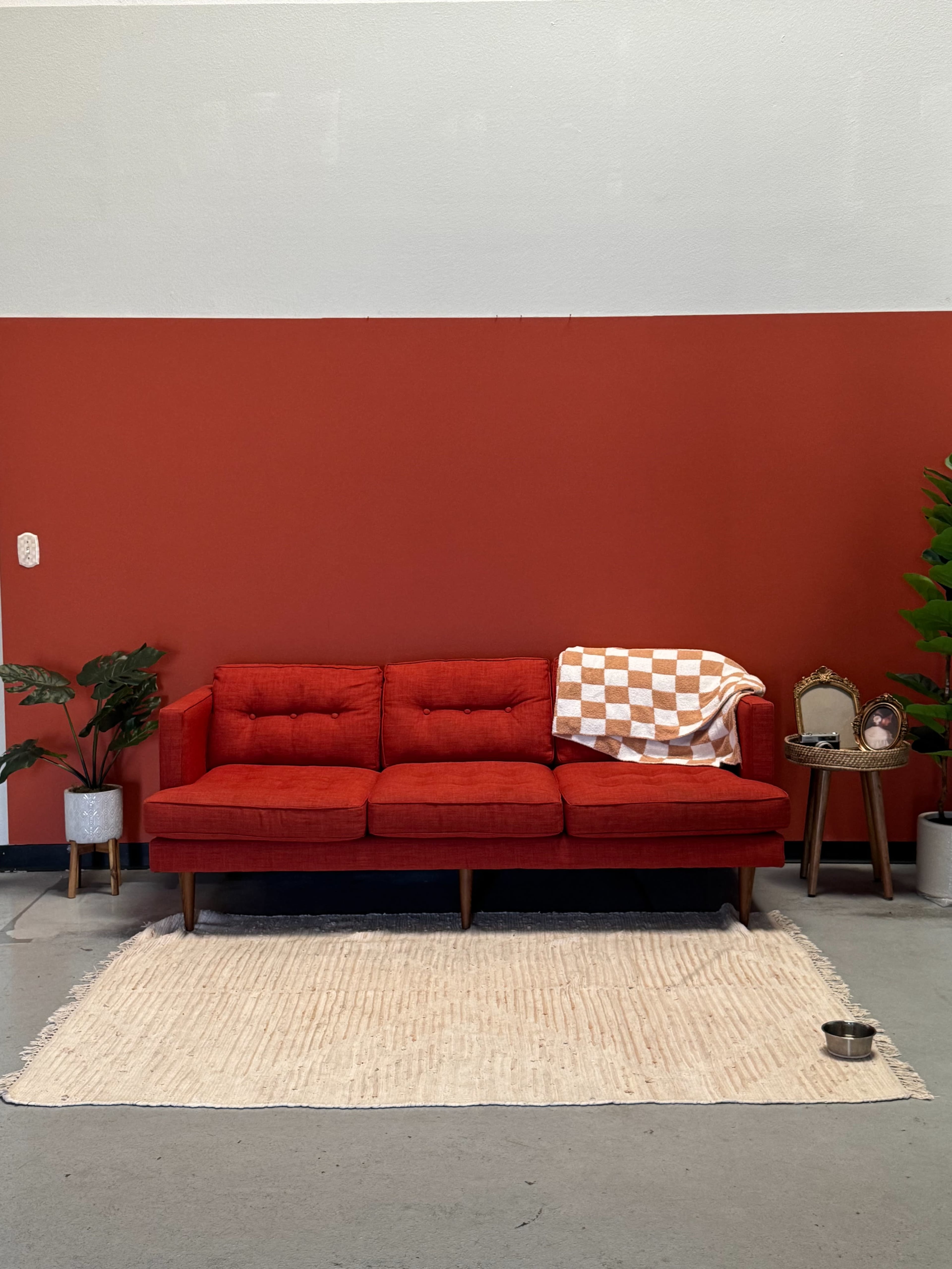 A red couch with tufted cushions is positioned against a two-tone red wall, accompanied by a woven rug, a small side table with a framed picture, and decorative plants.