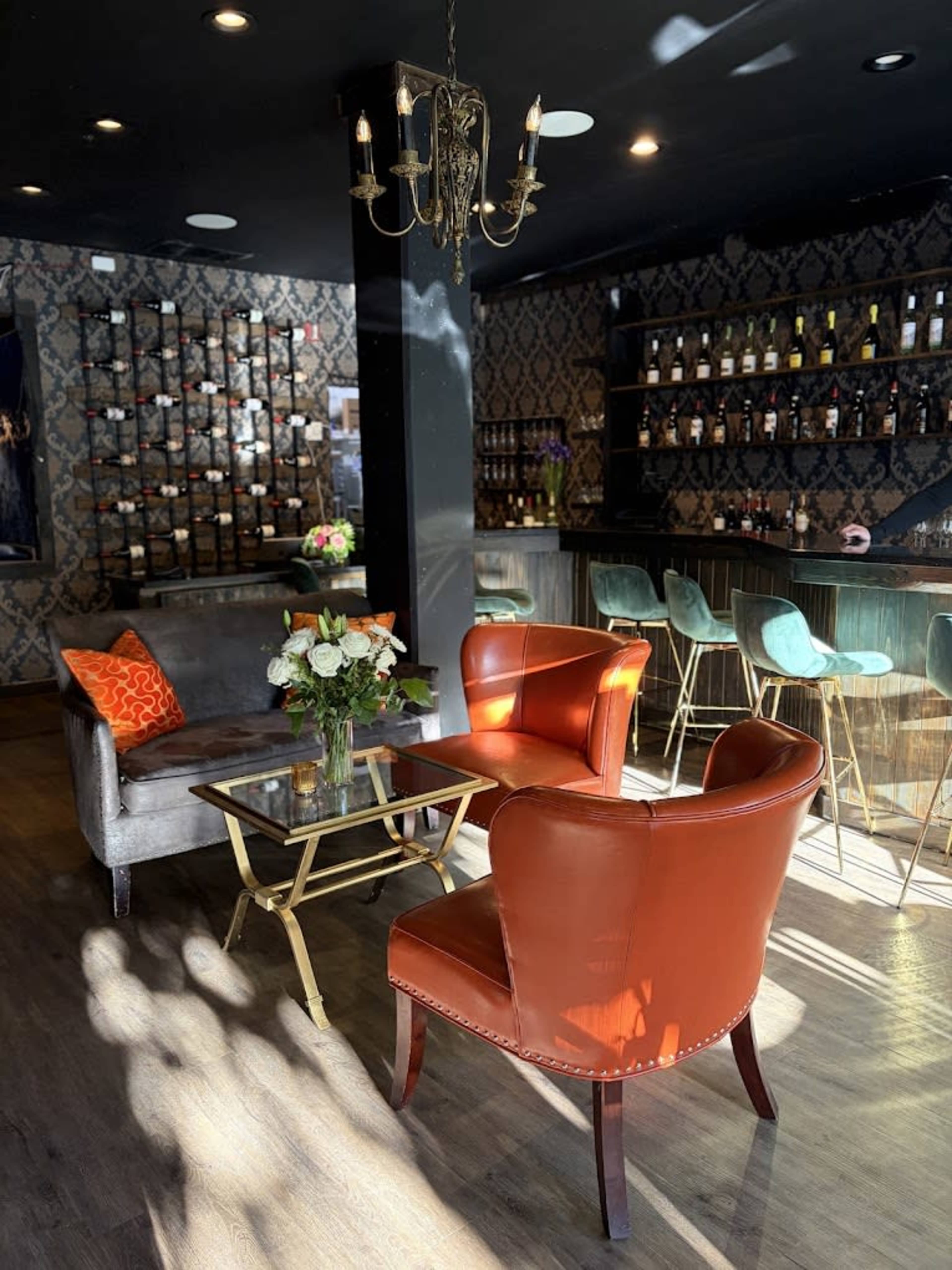 The image shows a stylish lounge area with a bar, featuring two orange chairs, a grey couch, a glass table, and a wall displaying various bottles.