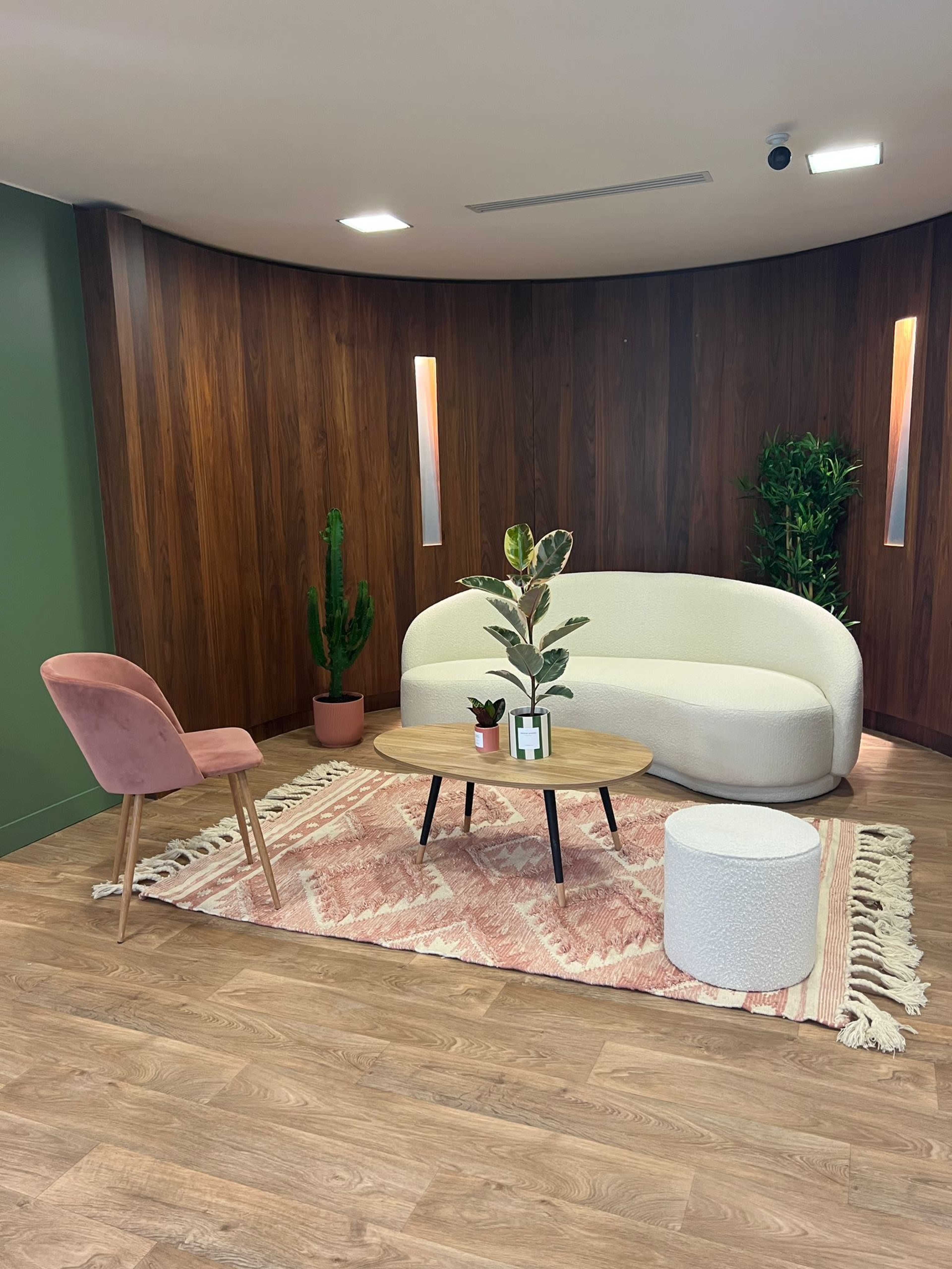 A modern sitting area features a curved white sofa, a wooden coffee table, a pink chair, and decorative plants on a patterned rug.