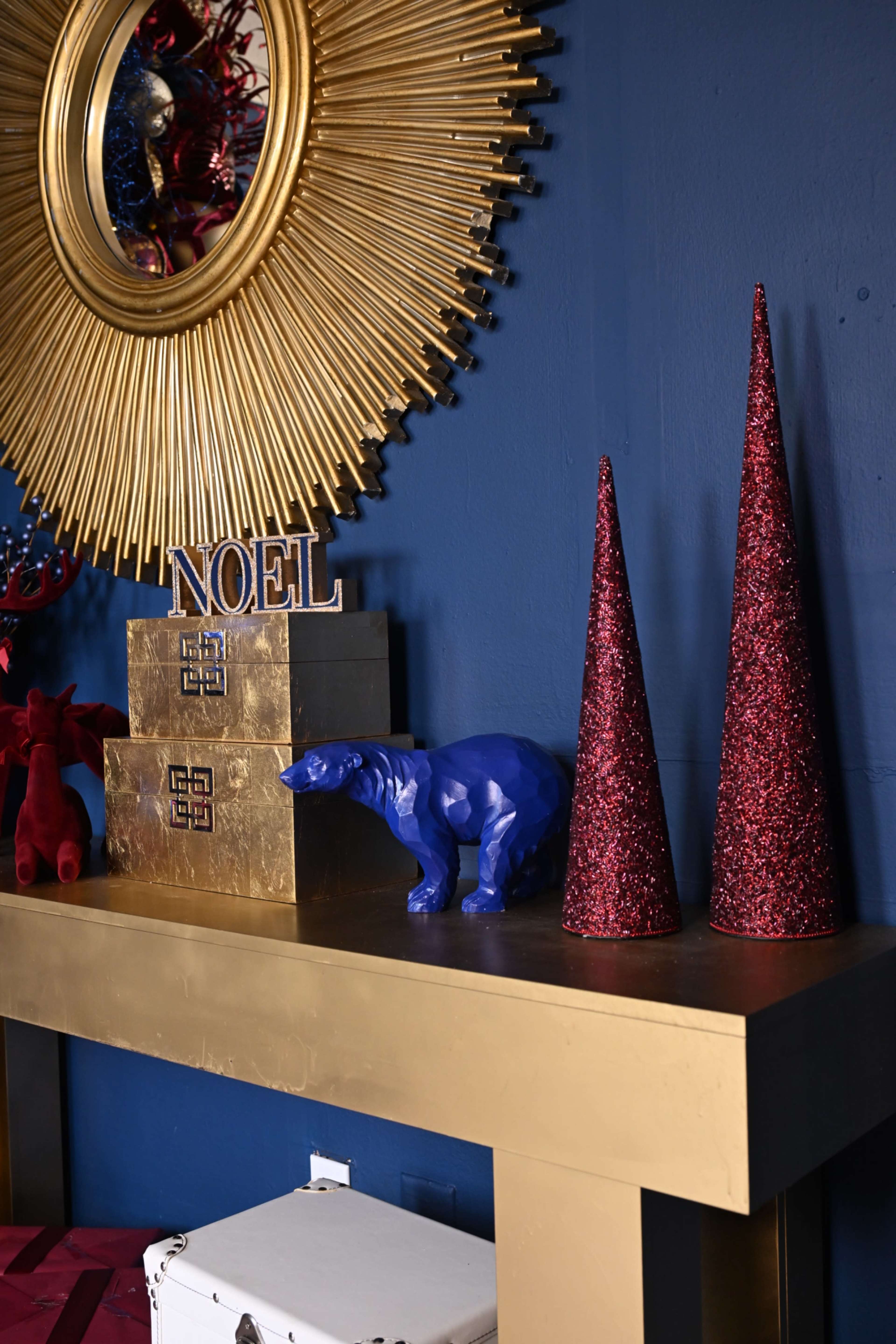 A gold table against a blue wall displays a mirror, a "NOEL" sign, two glittery red trees, a blue polar bear figurine, and gold boxes.