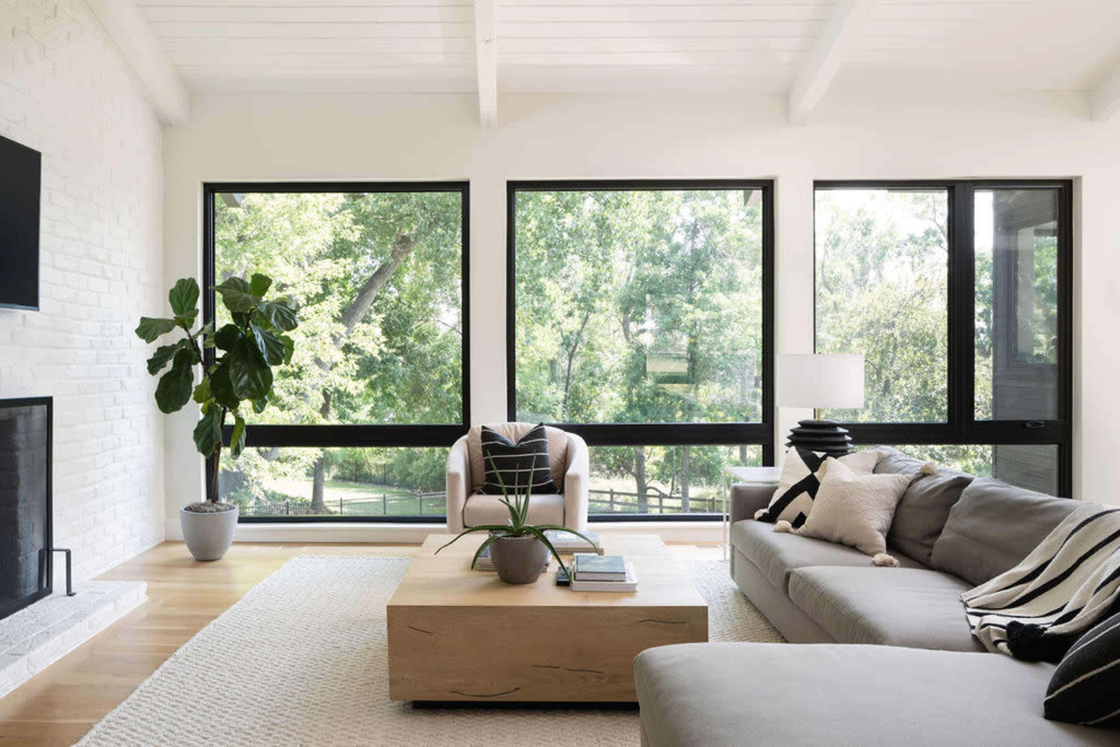 The image shows a modern living room featuring large windows that provide a view of greenery, a light-colored sofa, a coffee table, and a potted plant.
