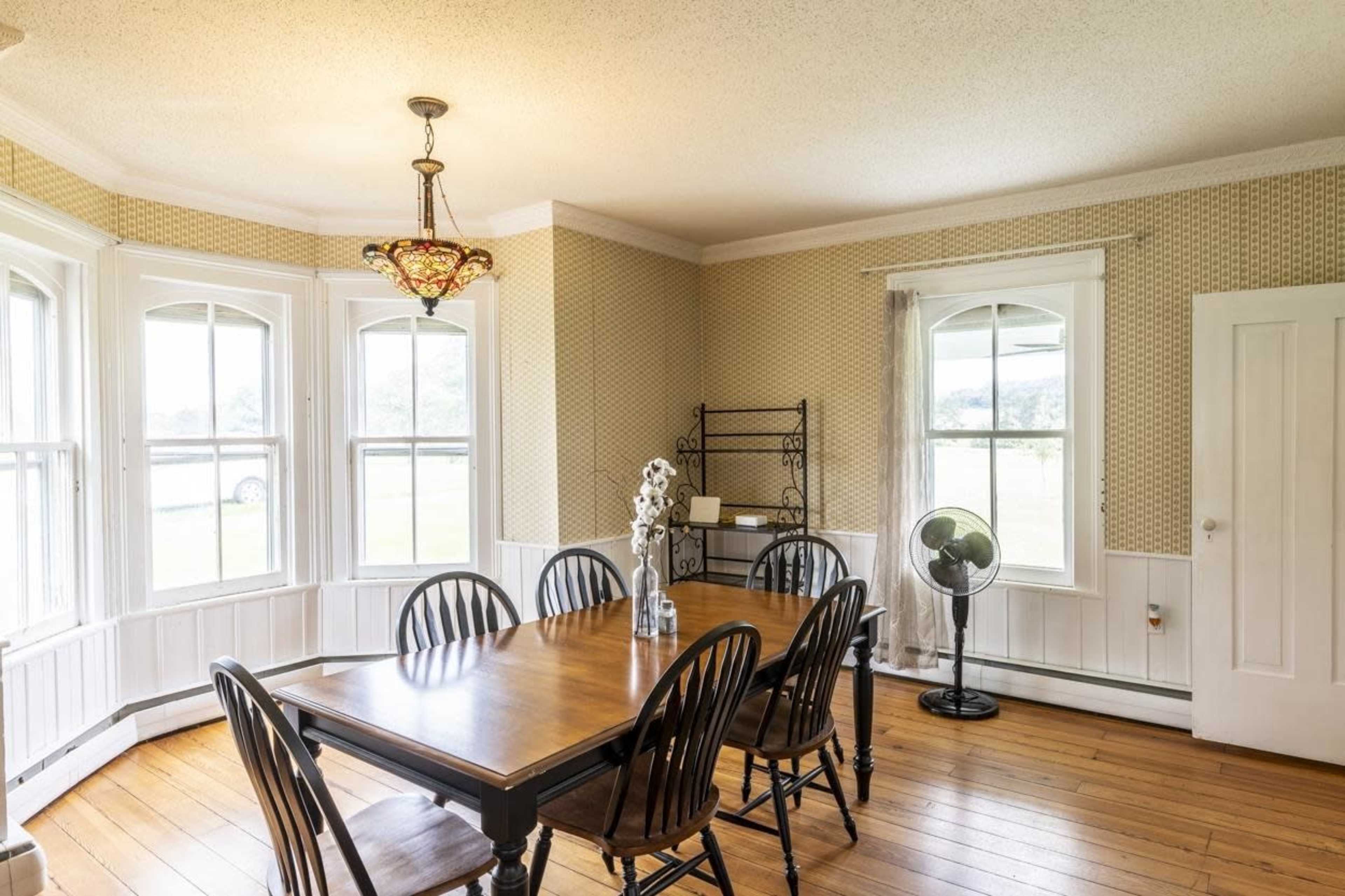 A dining room features a wooden table surrounded by black chairs, large windows allowing natural light, and a chandelier hanging from the ceiling.